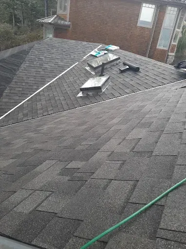 A dark shingle roof with skylights, a green hose, and a brick house in the background.