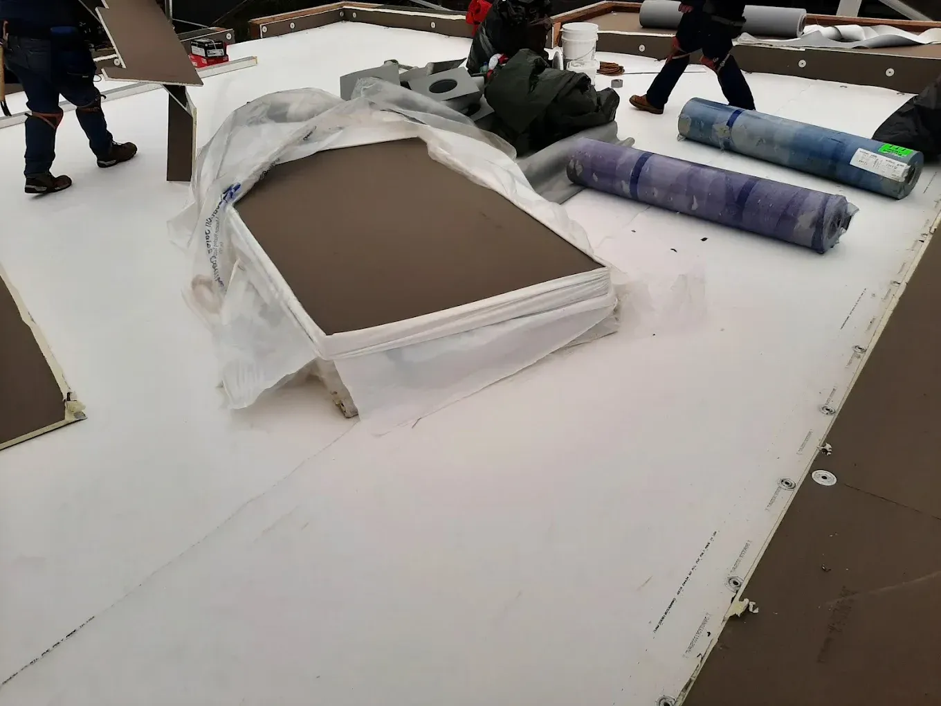 Workers installing a white roof. Stacked materials covered in plastic, rolls of roofing, and people walking.