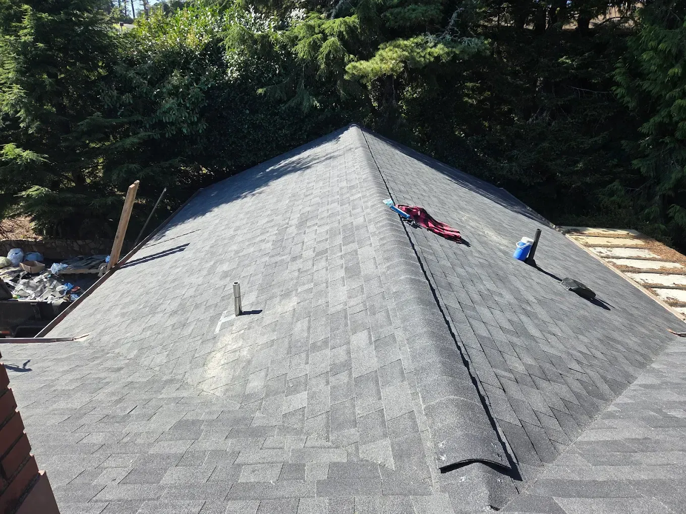 Roof with dark shingles, a few tools, and trees in the background on a sunny day.