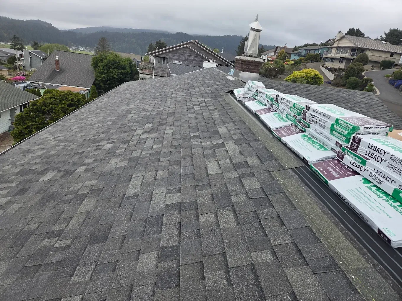 Roof with stacked shingle bundles, overcast day. Buildings and landscape visible in background.