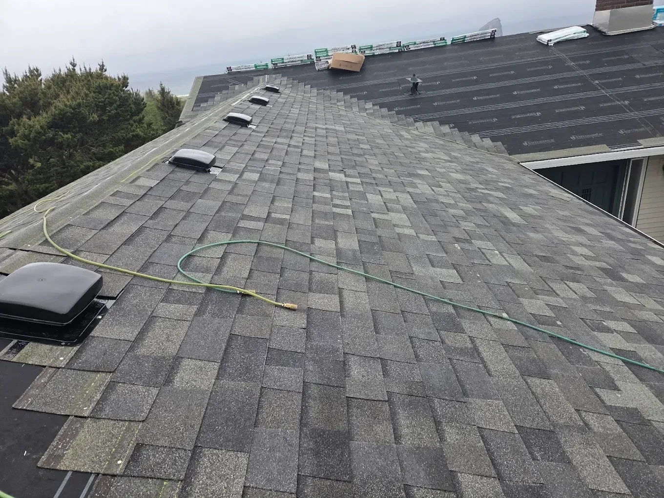 Gray asphalt shingle roof with several black vents, a green hose, and a partly cloudy sky.