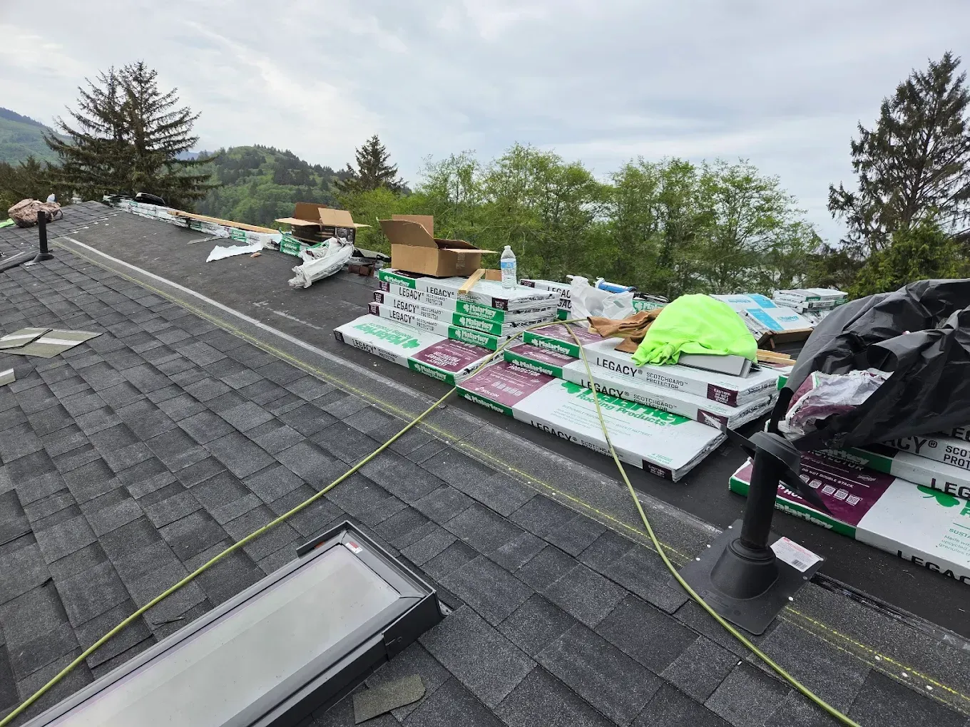 Roof with bundles of shingles, boxes, and safety equipment; overcast day.