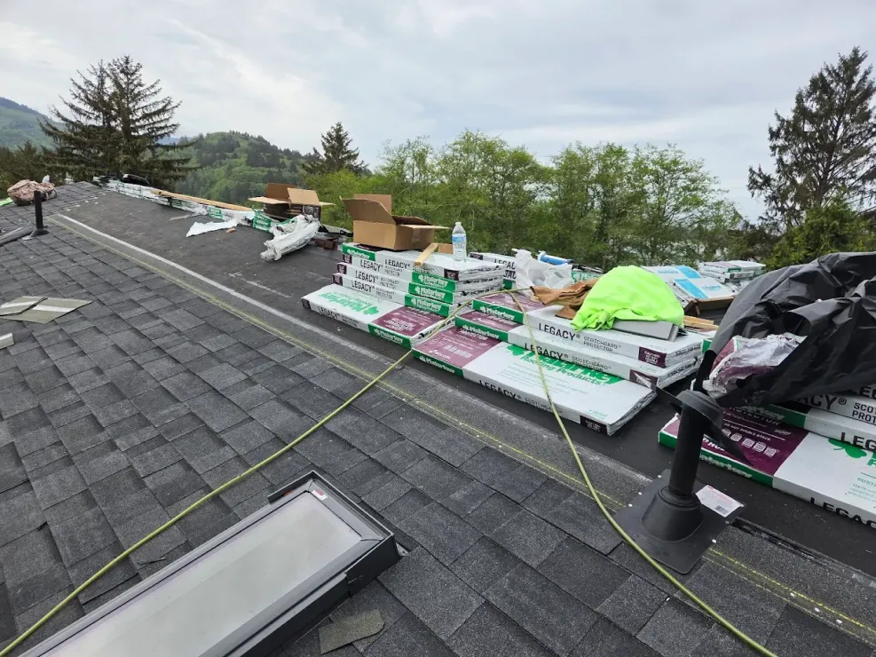 Roof with stacks of new shingles and equipment on a cloudy day, trees in the background.