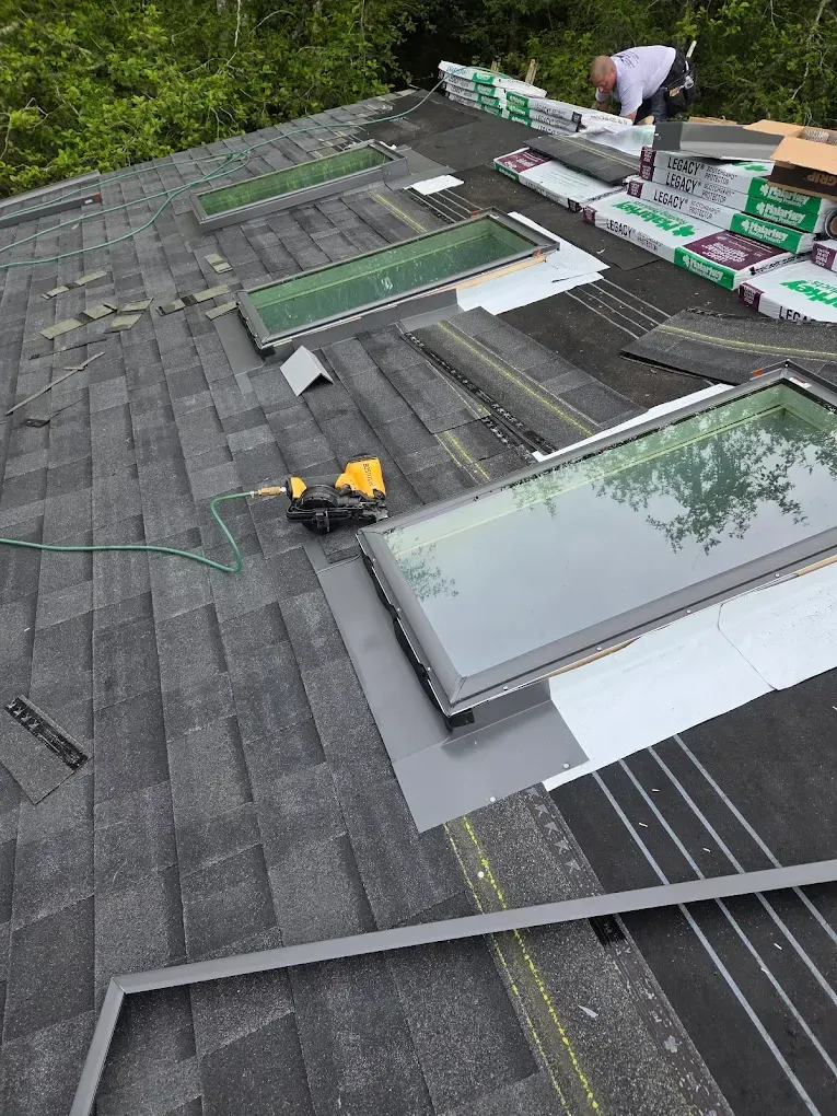 Roofer installing shingles around skylights on a gray asphalt shingle roof.