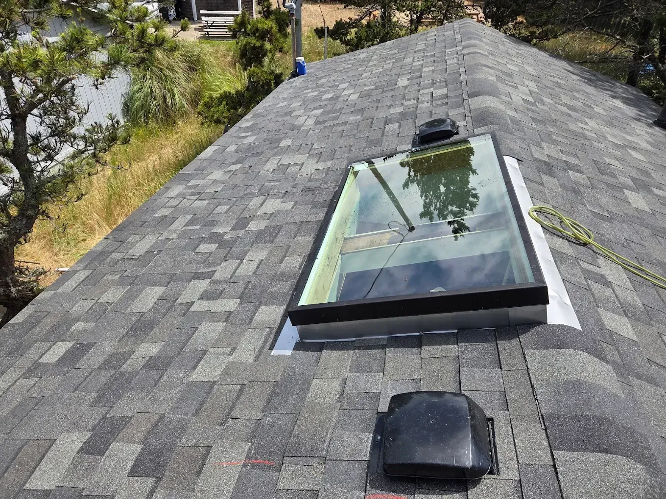 Gray shingle roof with a skylight and vent. Viewed from above with a rope and trees in the background.