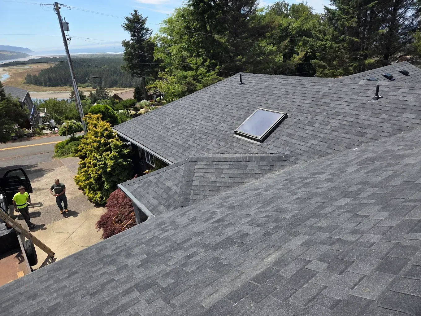 View of a house with a dark gray shingle roof, two people, a car, and a scenic background.