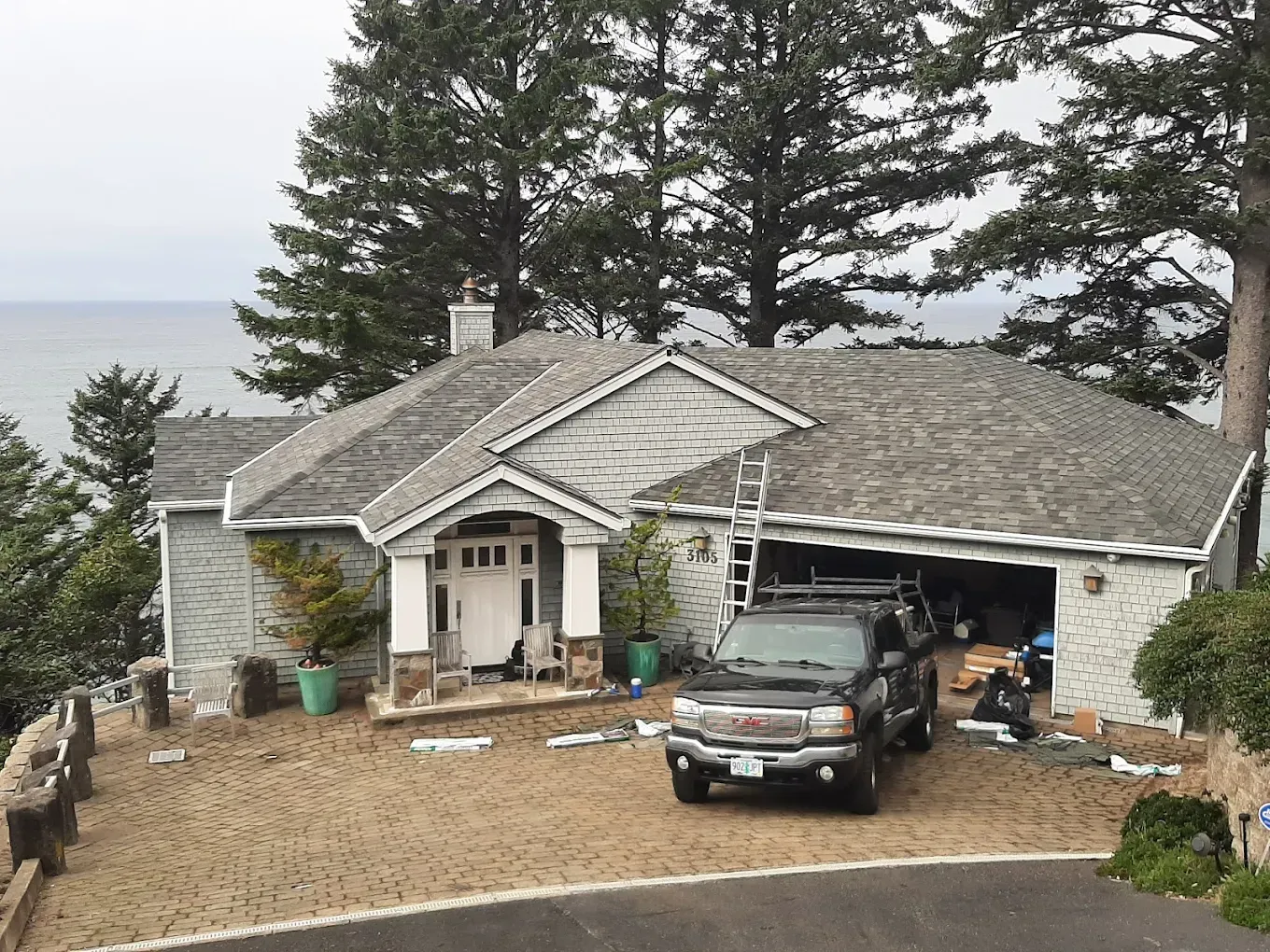 House with dark truck in driveway, near ocean. Grey roof, cloudy sky.