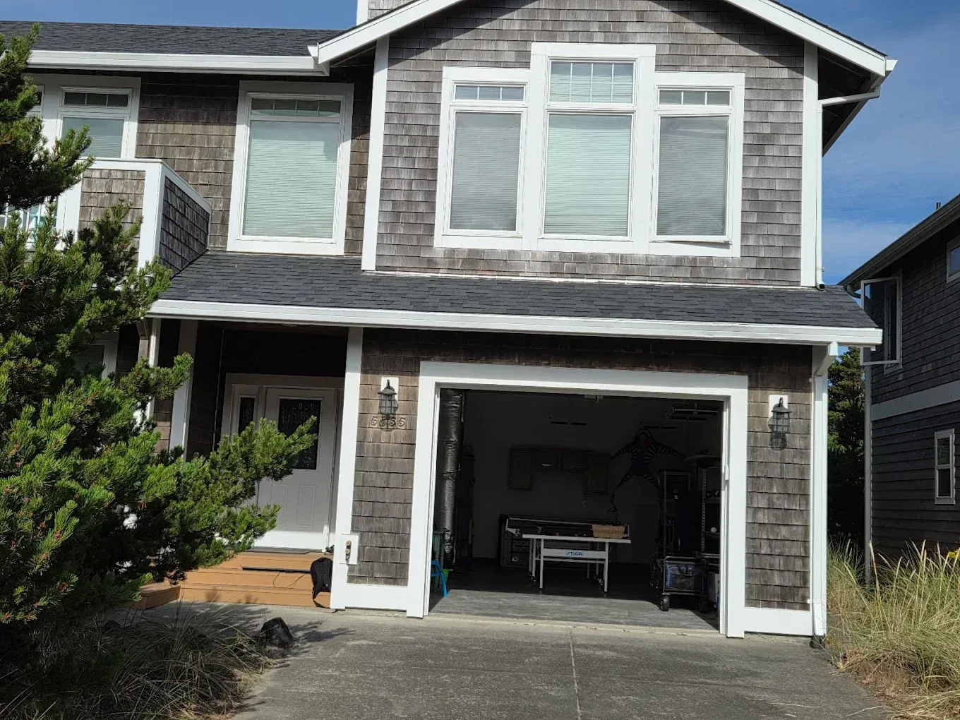 Two-story house with weathered shingles, open garage, and gray driveway; sunny day.