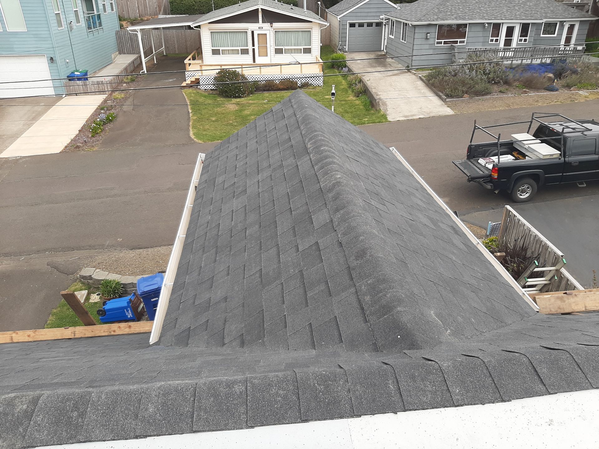 View of a dark shingled roof with houses and a truck in the background on a sunny day.