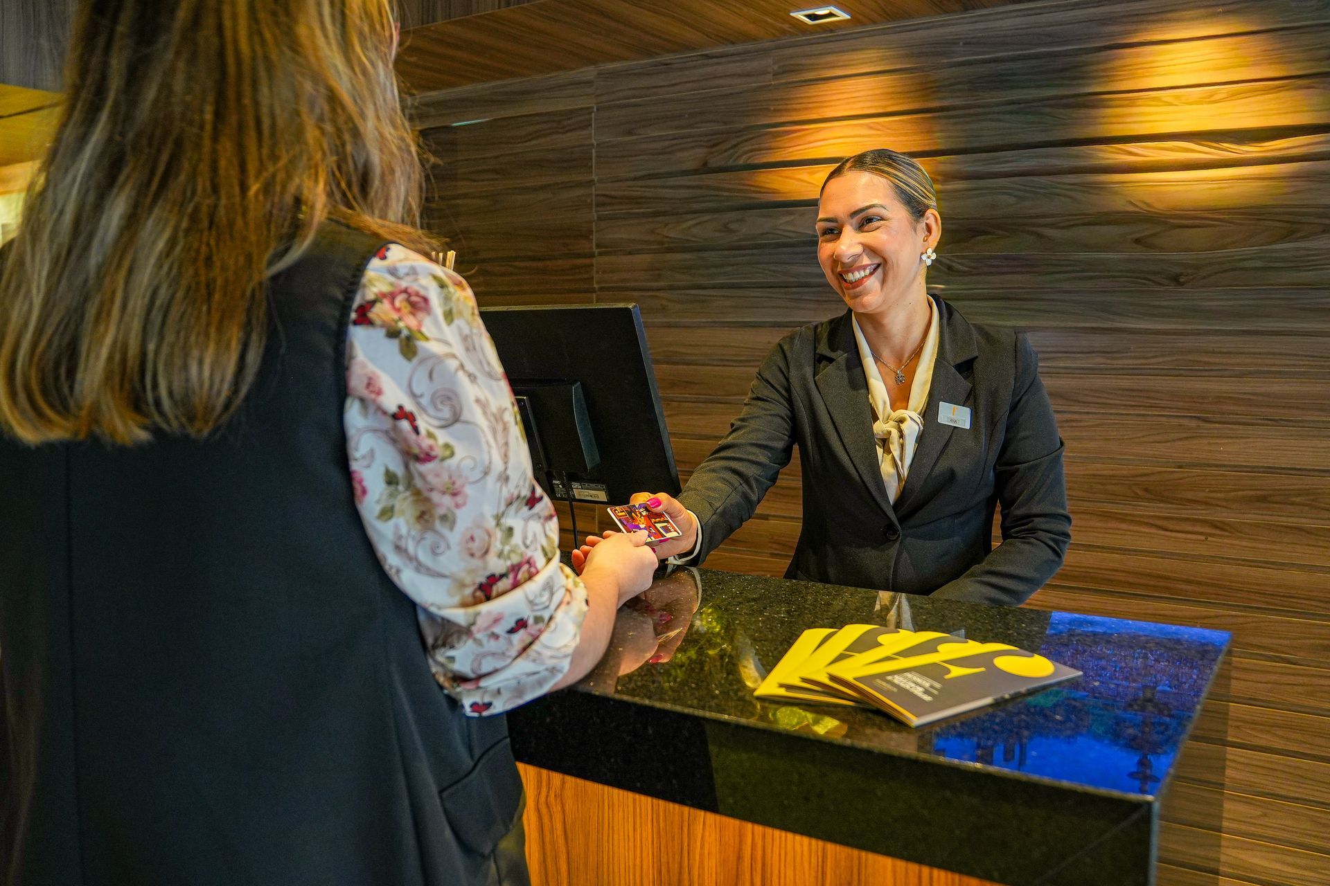Uma recepcionista sorri enquanto entrega um cartão-chave a um hóspede na recepção do hotel.