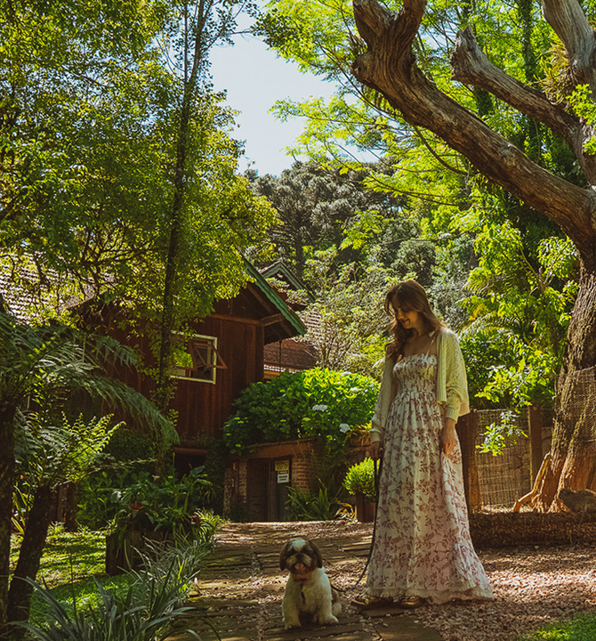 Uma pessoa vestida com um vestido florido e um casaquinho leve passeia com um cachorrinho fofo por uma trilha de pedra em um jardim ensolarado e arborizado.