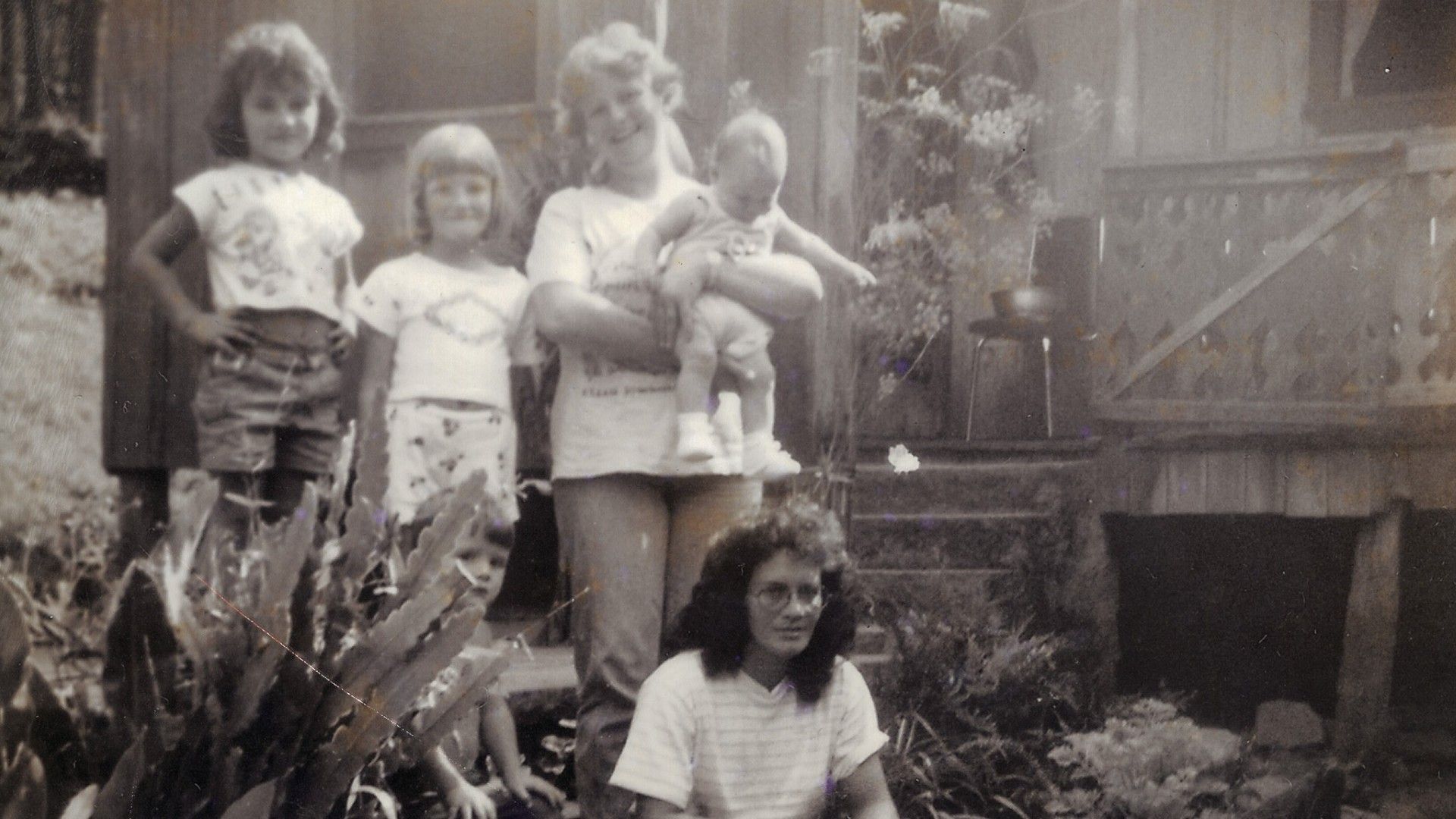 Uma fotografia antiga em preto e branco de um grupo posando em frente a uma casa, com uma mulher segurando um bebê e outras pessoas em pé.