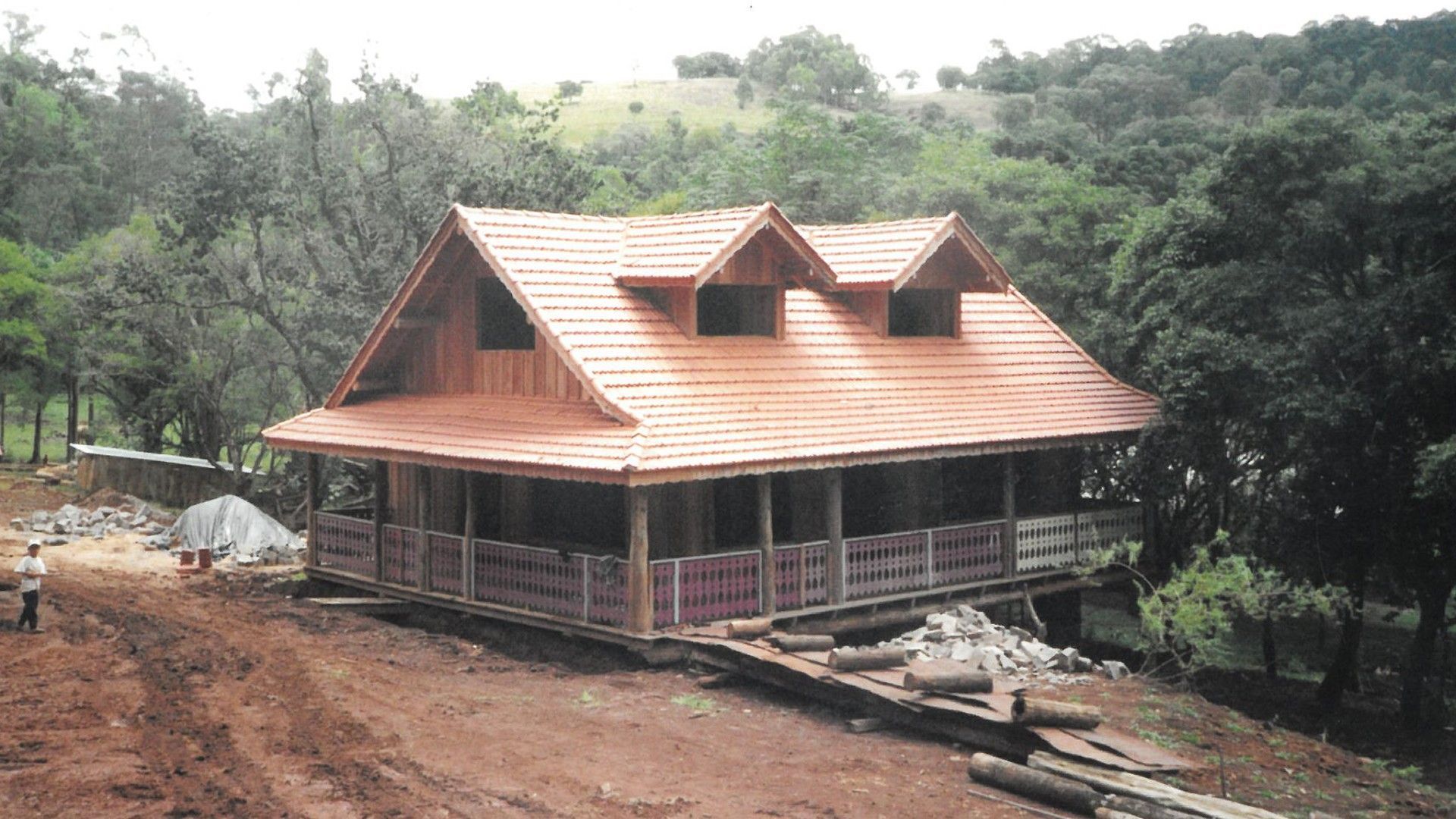 Uma cabana rústica de madeira em construção, com telhado de duas águas e duas janelas tipo mansarda, situada em uma paisagem arborizada.