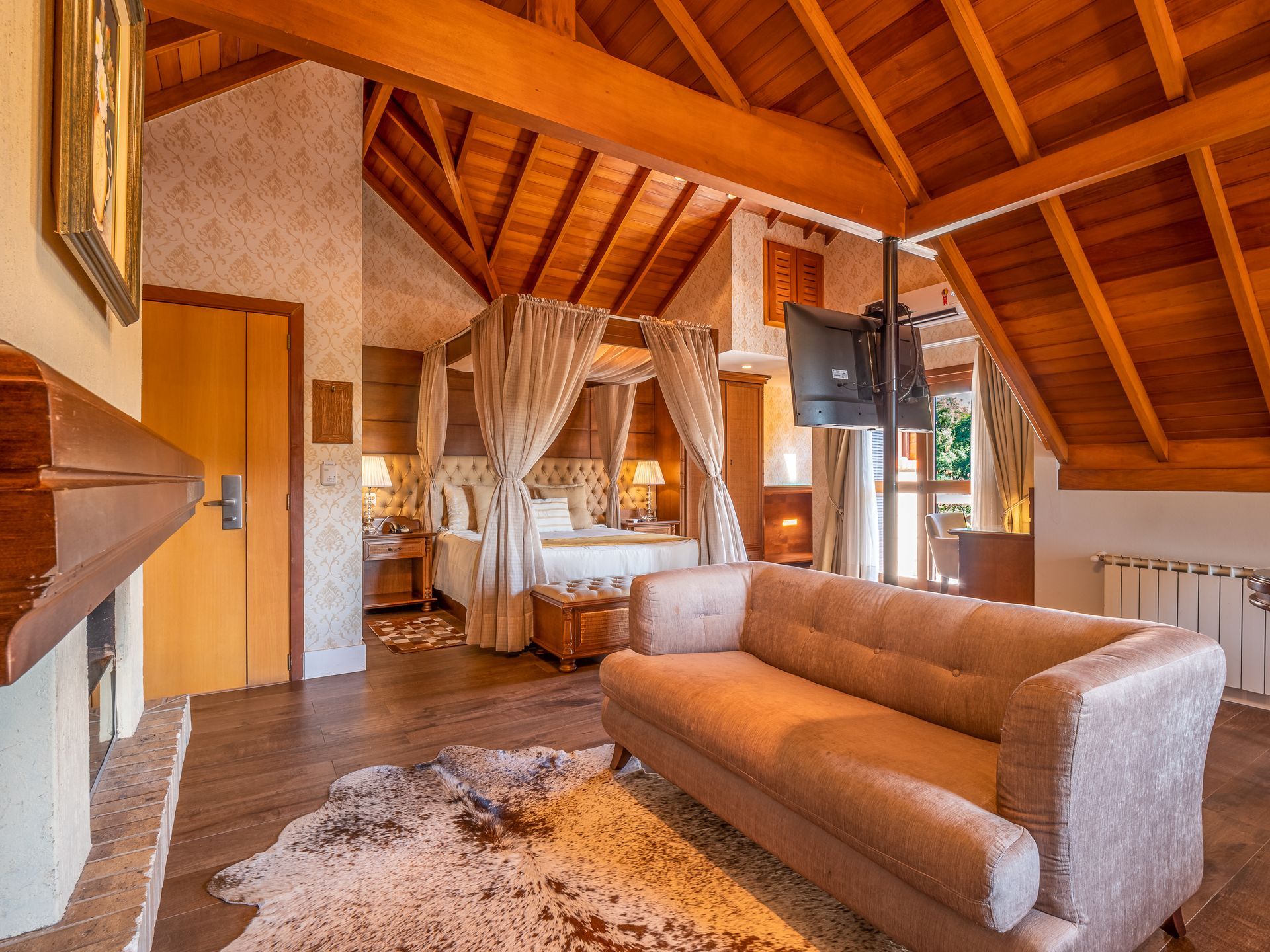 Uma suíte de hotel aconchegante, com painéis de madeira, sofá, cama com cortinas e lareira em primeiro plano.