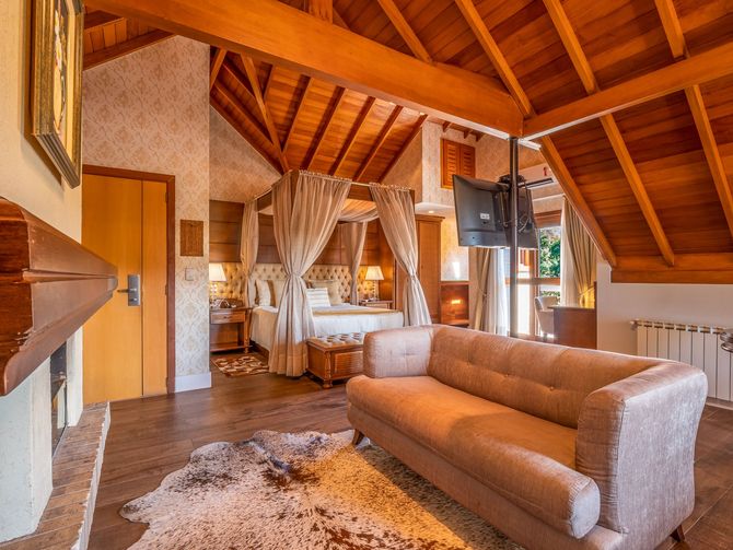 Uma suíte de hotel aconchegante, com painéis de madeira, sofá, cama com cortinas e lareira em primeiro plano.