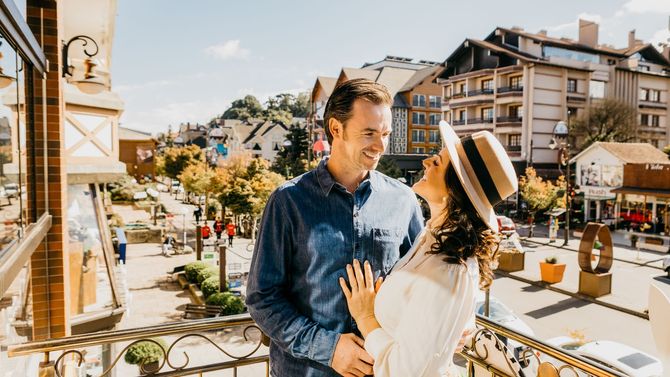 Um casal com roupas casuais rindo junto em uma varanda ensolarada com vista para uma praça de estilo europeu.