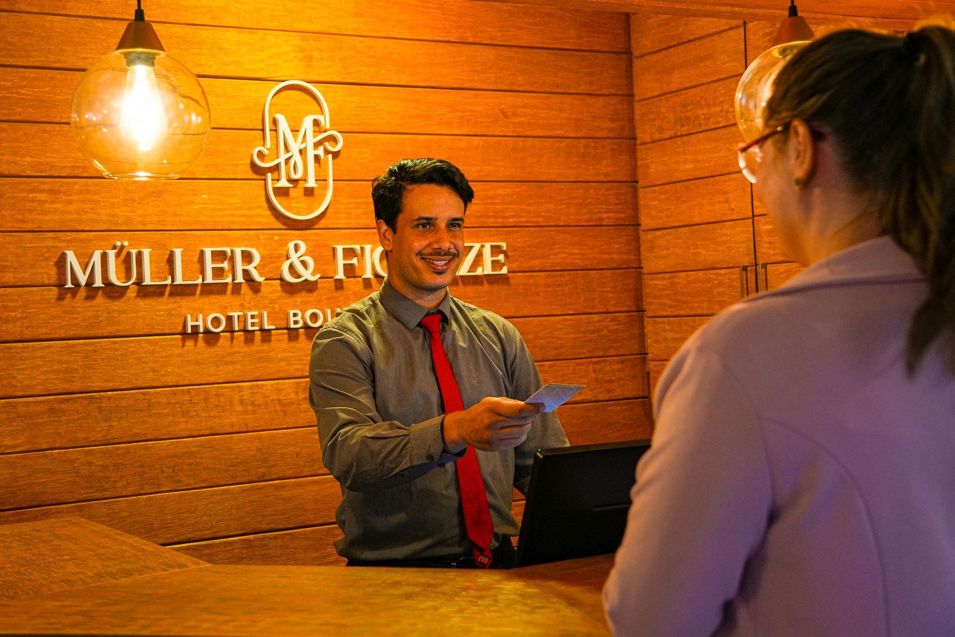 Uma recepcionista de hotel, vestindo camisa cinza e gravata vermelha, entrega um documento a um hóspede em um balcão de madeira no saguão.