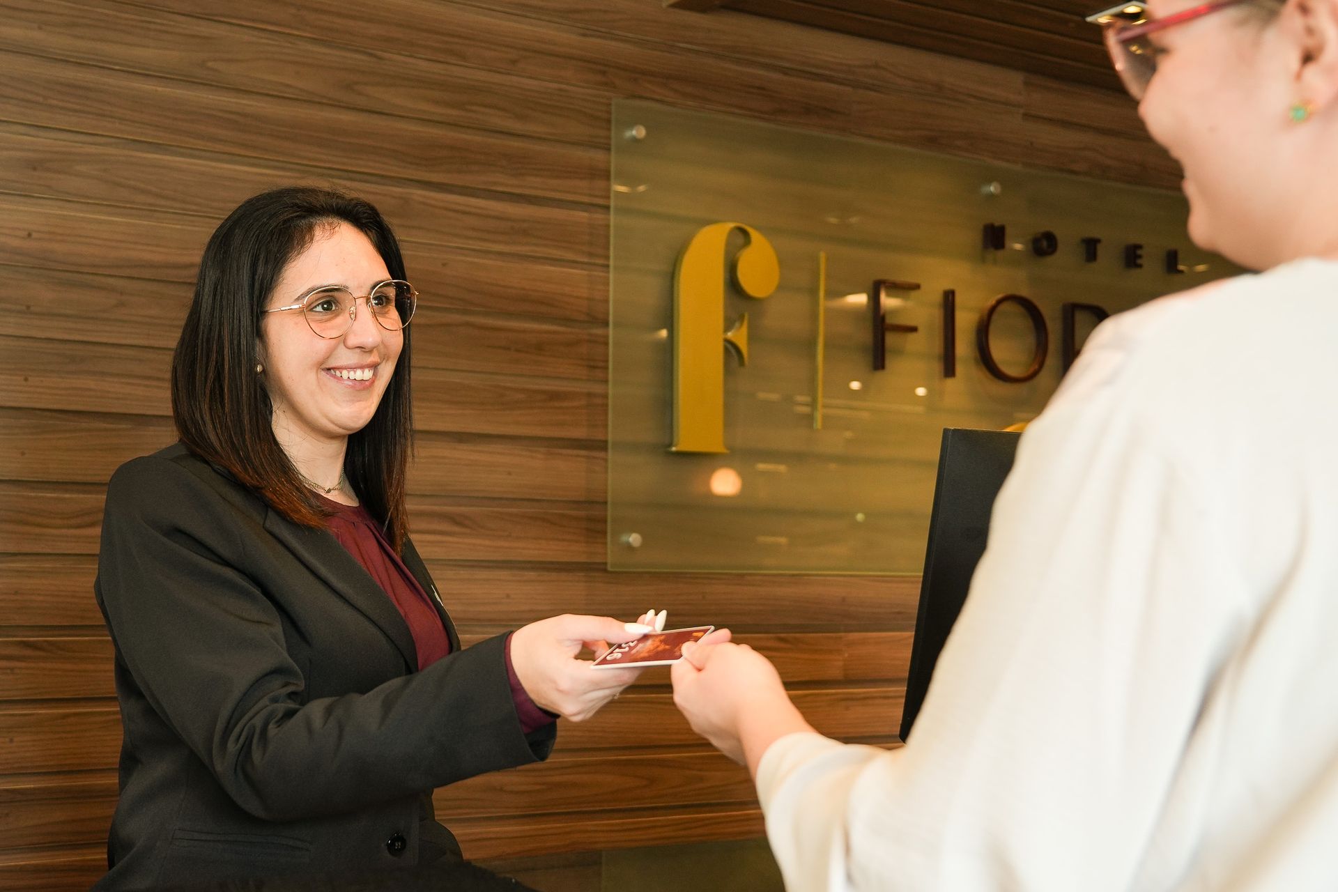 Um funcionário sorridente, de terno e gravata, entrega o cartão-chave do hotel a um hóspede em um balcão de recepção com painéis de madeira.