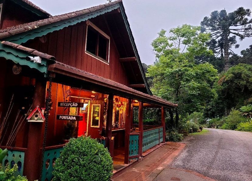 Uma casa de campo rústica, de madeira vermelho-escura, com uma varanda com detalhes em verde e iluminação interior aconchegante, situada ao lado de uma estrada de terra.