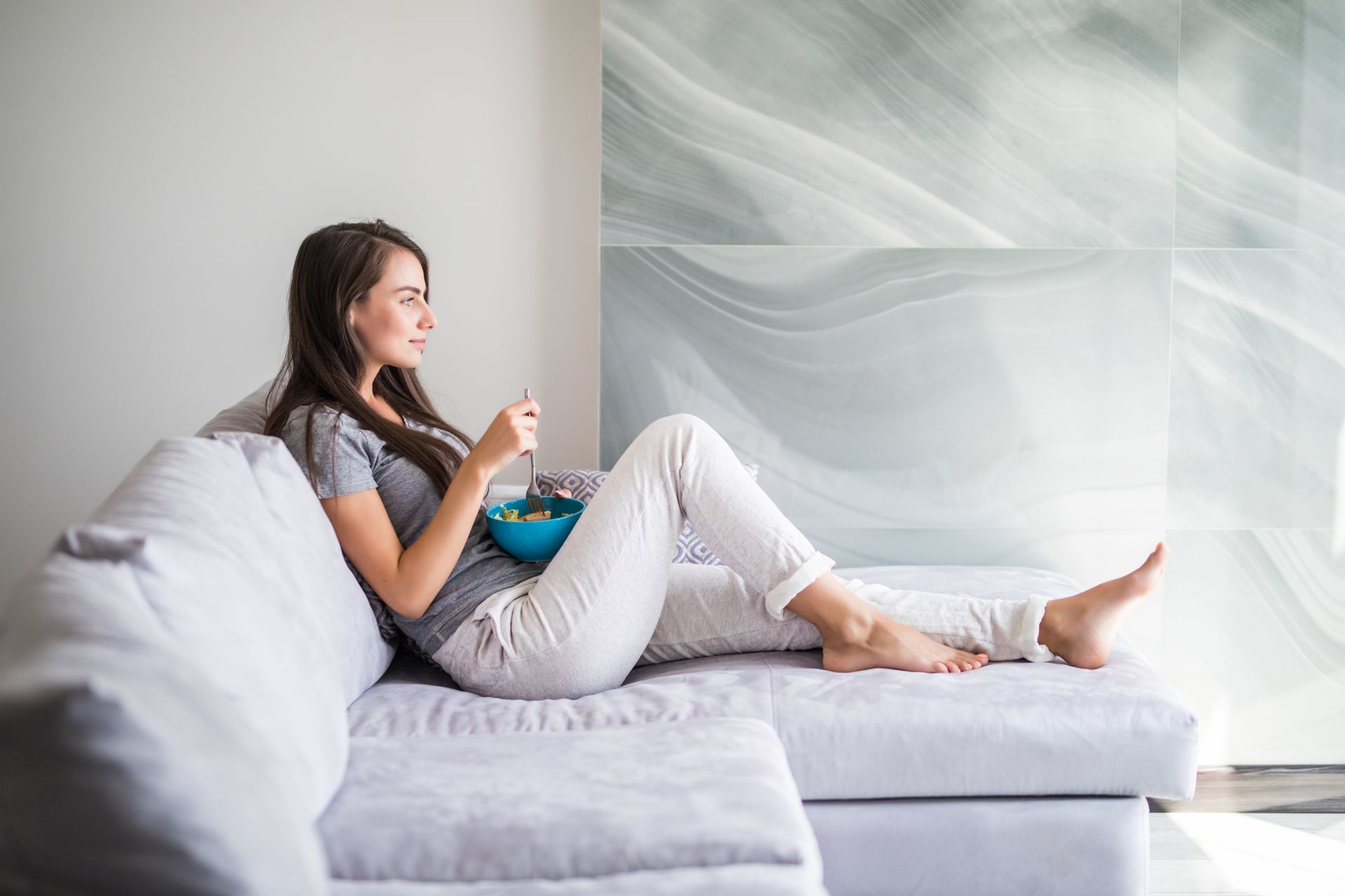 A person sits on a light gray sofa, relaxing and eating from a small blue bowl in a minimalist, light-filled room.