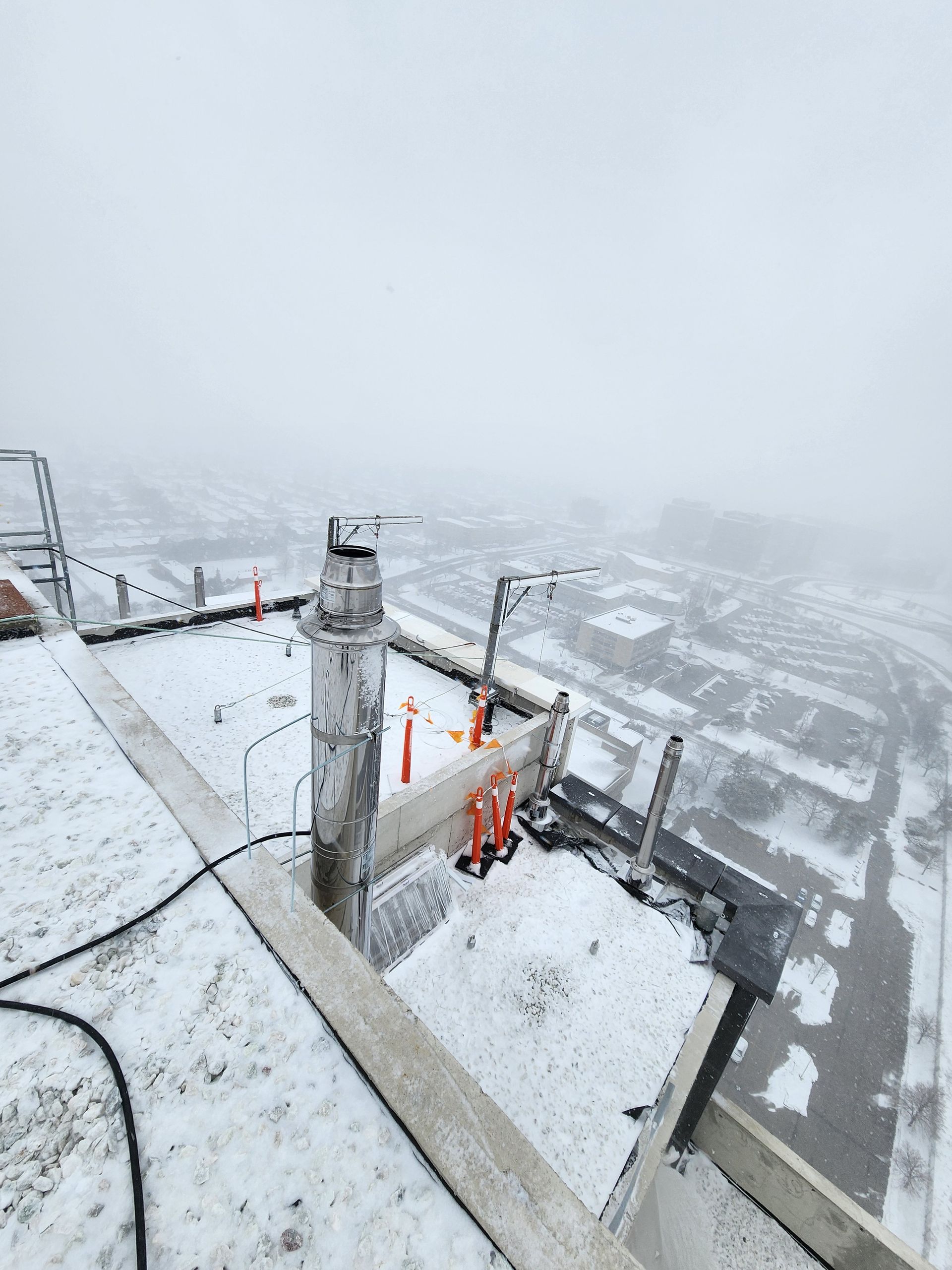 Snowy rooftop with a vent stack and orange construction cones. Foggy cityscape in the background.
