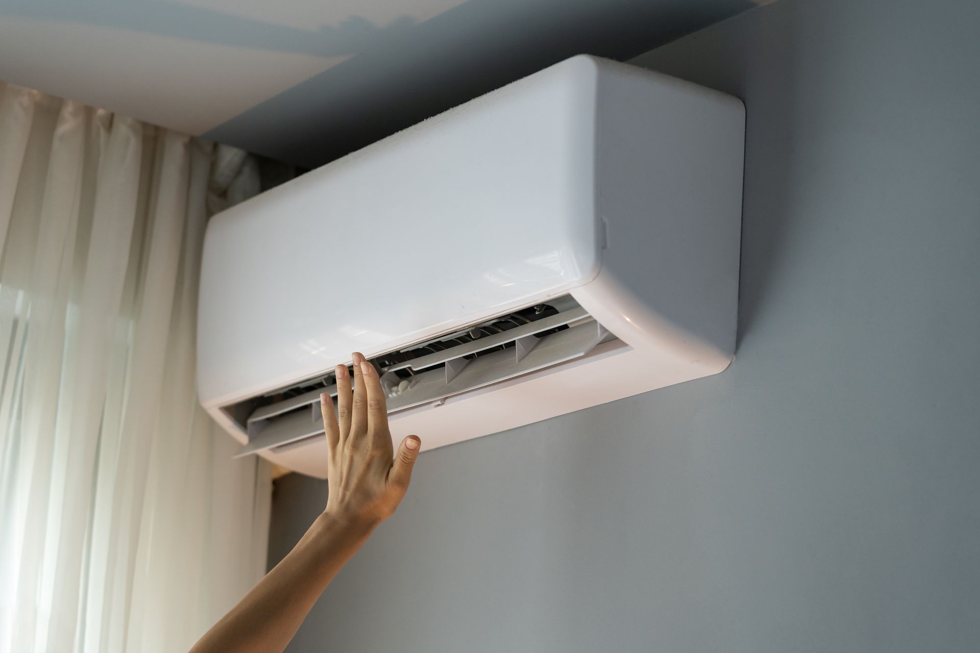 A hand reaches toward a white wall-mounted air conditioning unit in a room with light curtains.