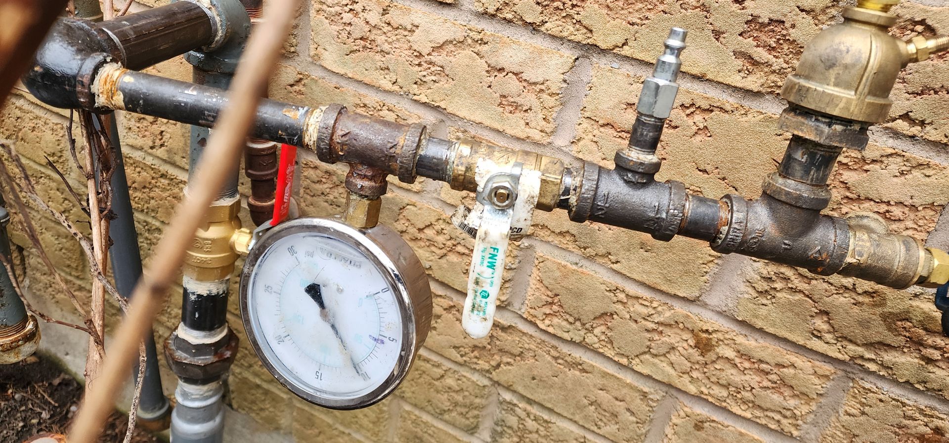Pipes with a pressure gauge, valve, and fittings against a brick wall.