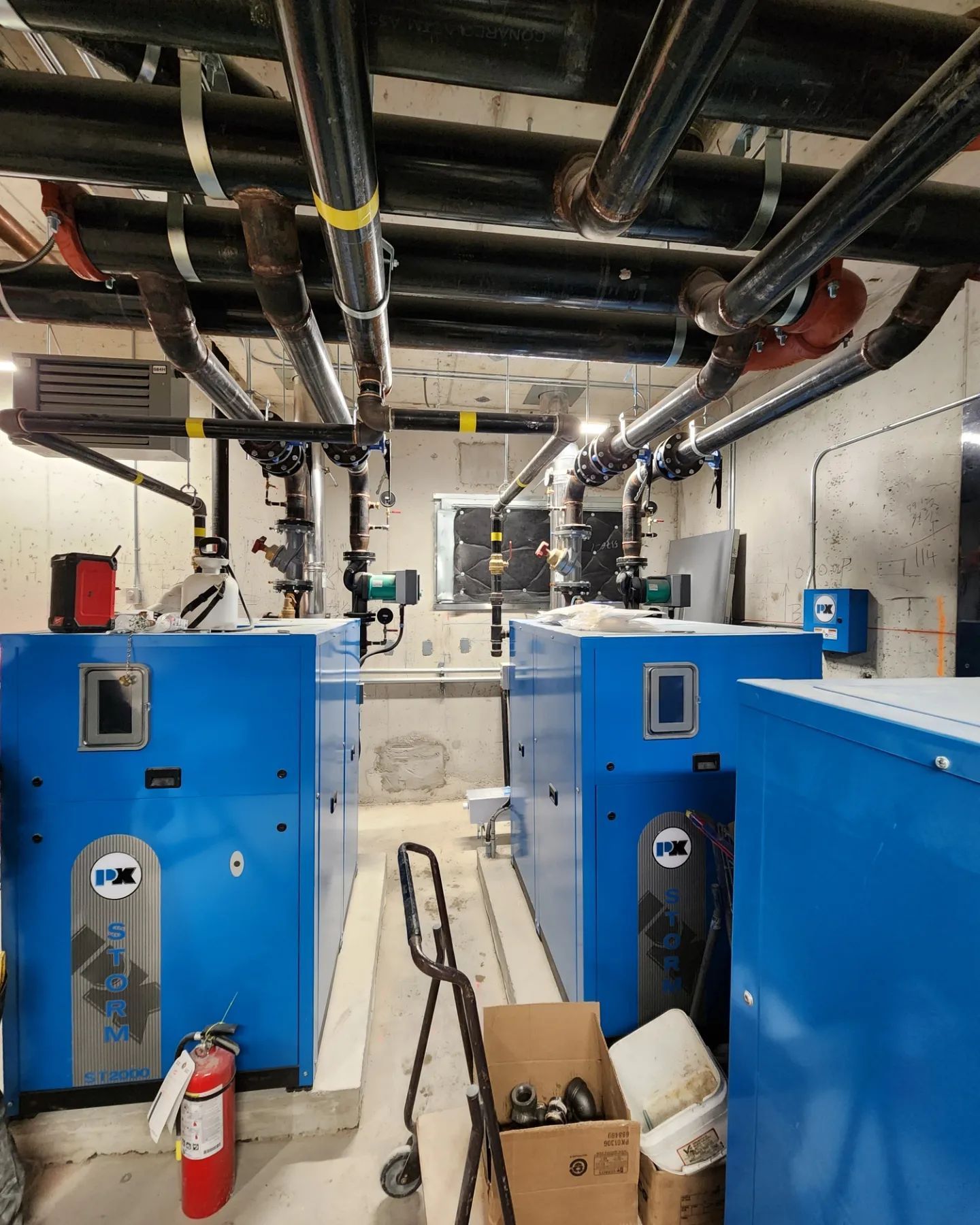 Blue industrial heating units in a utility room with black pipes overhead. A fire extinguisher and small cart are visible.