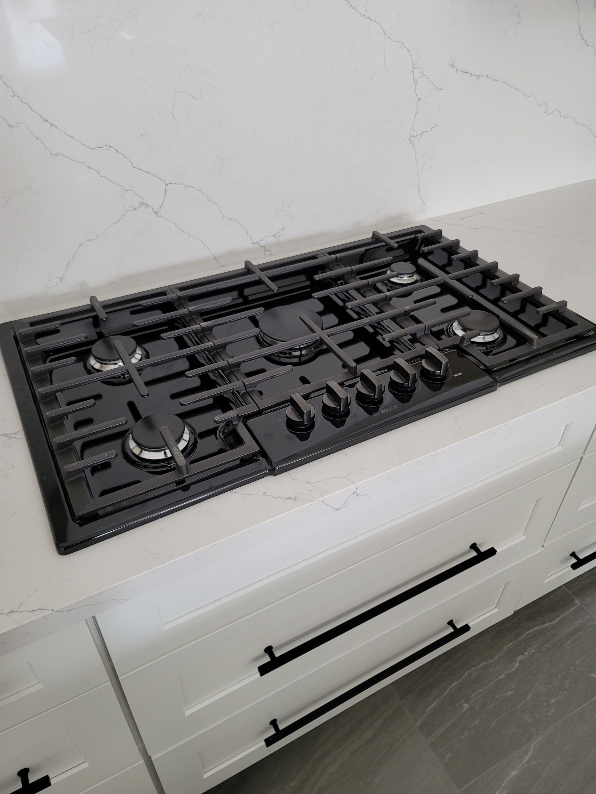 Black gas cooktop on white countertop above white cabinets.