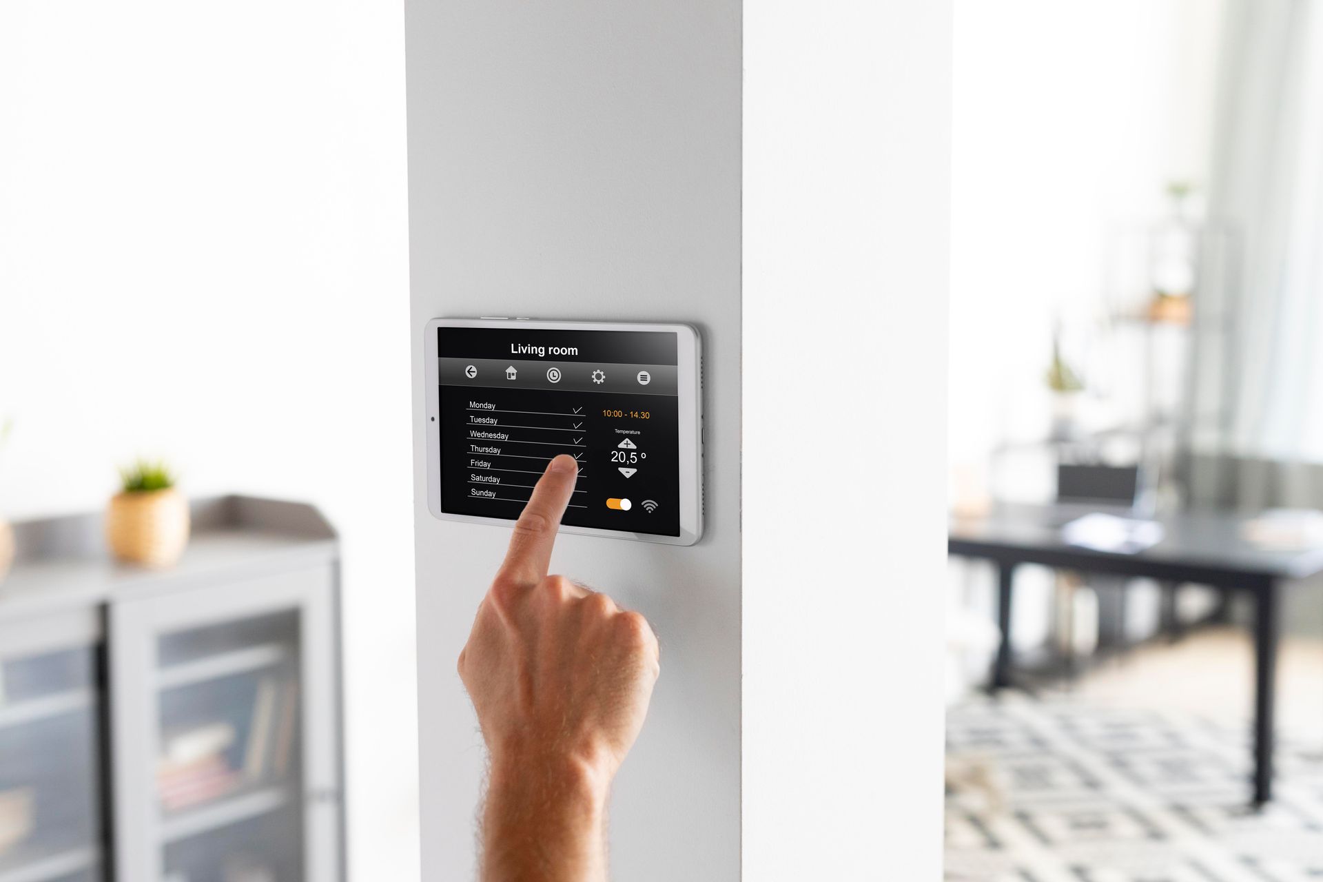 A person uses their finger to control a wall-mounted smart home display in a modern, bright interior setting.