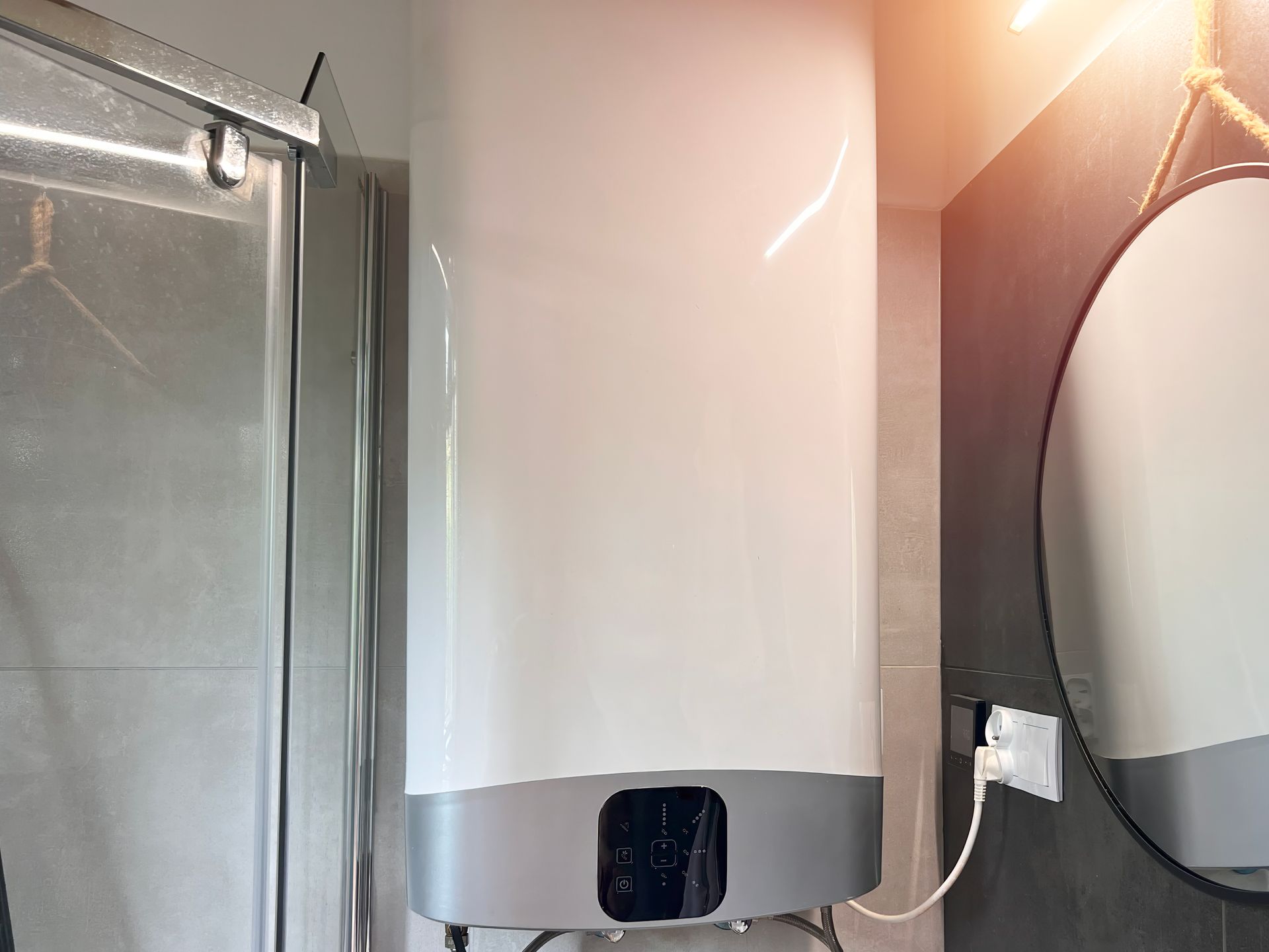 A white electric water heater with a digital display panel installed on a bathroom wall near a shower and a round mirror.