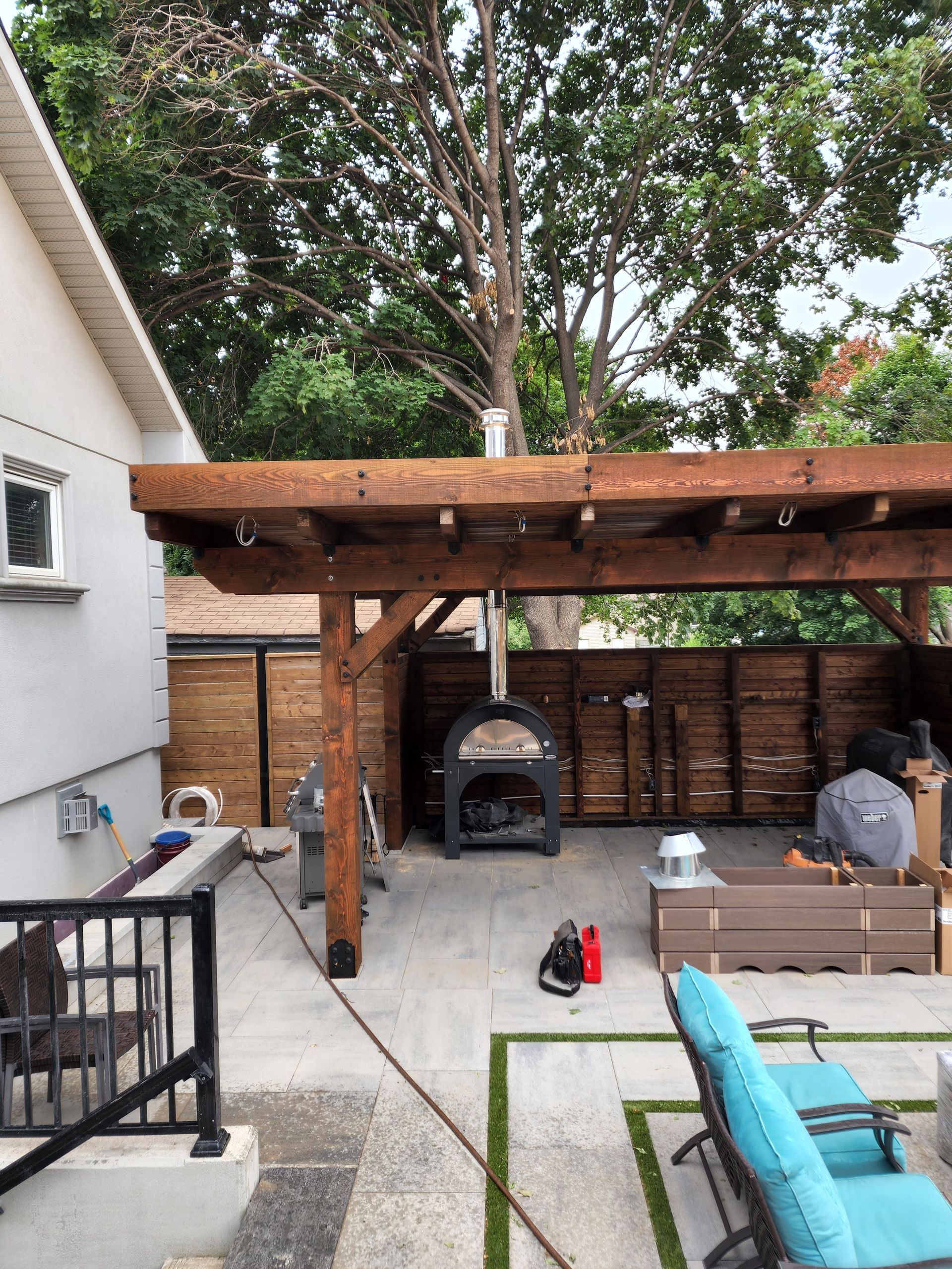 Outdoor patio with pizza oven under a wooden pergola; a person sits nearby.