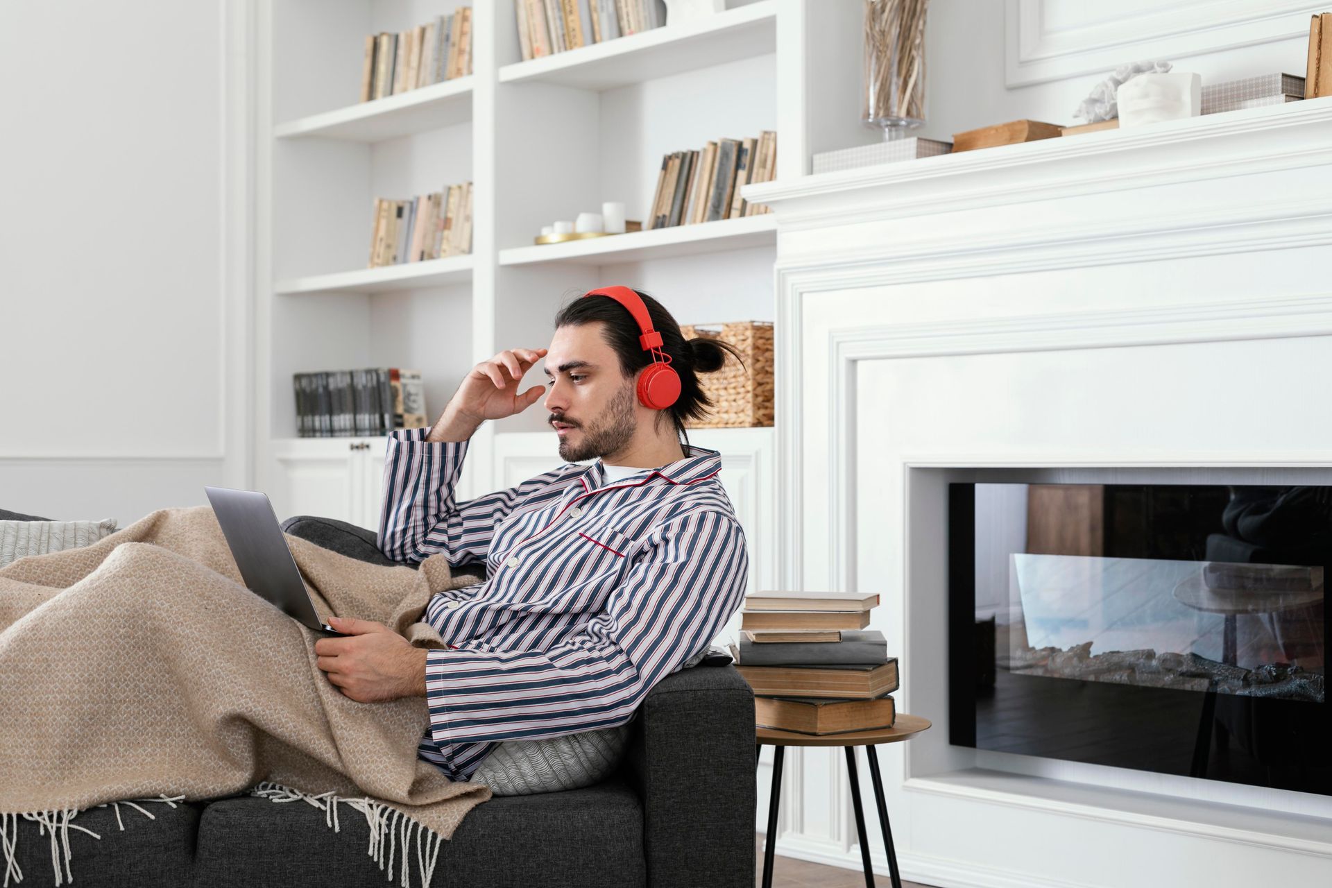 A person in striped pajamas and red headphones sits on a couch under a blanket, looking at a laptop in a bright room.