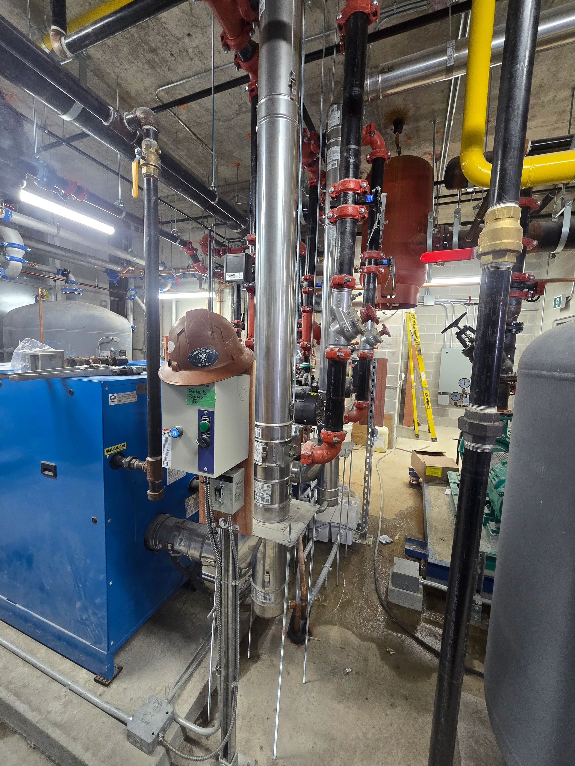 A boiler room with pipes, a blue machine, and a brown hard hat. Water is leaking on the floor.