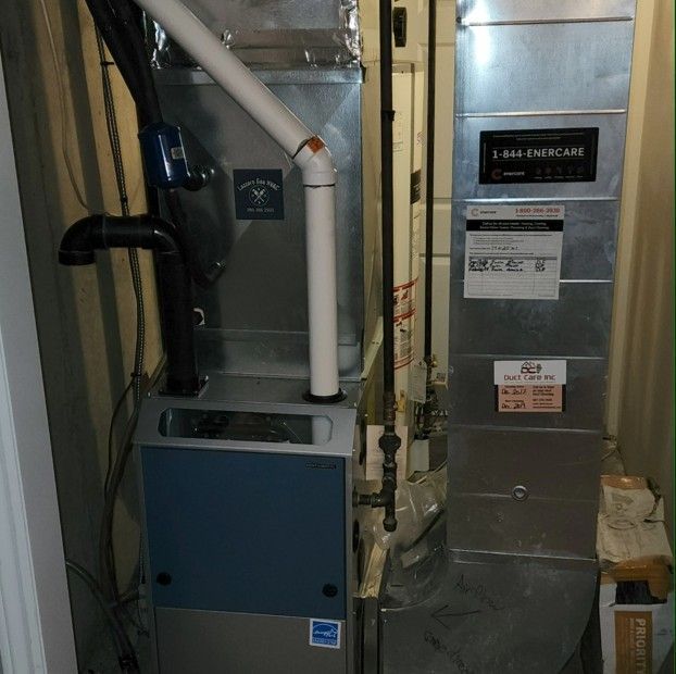 A furnace with metal ducts, white pipes, and a blue front panel inside a utility room.