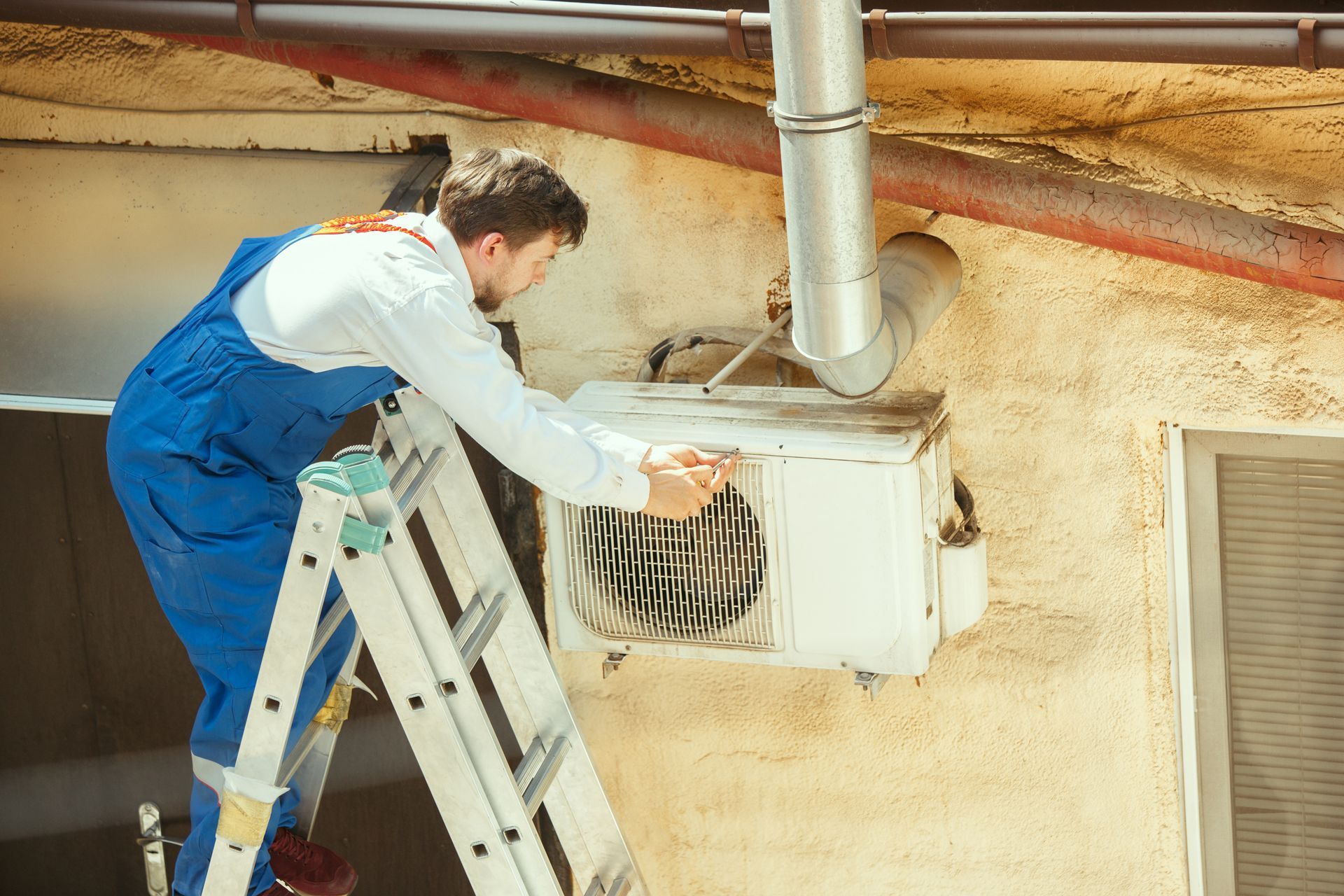 A man is standing on a ladder fixing an air conditioner.