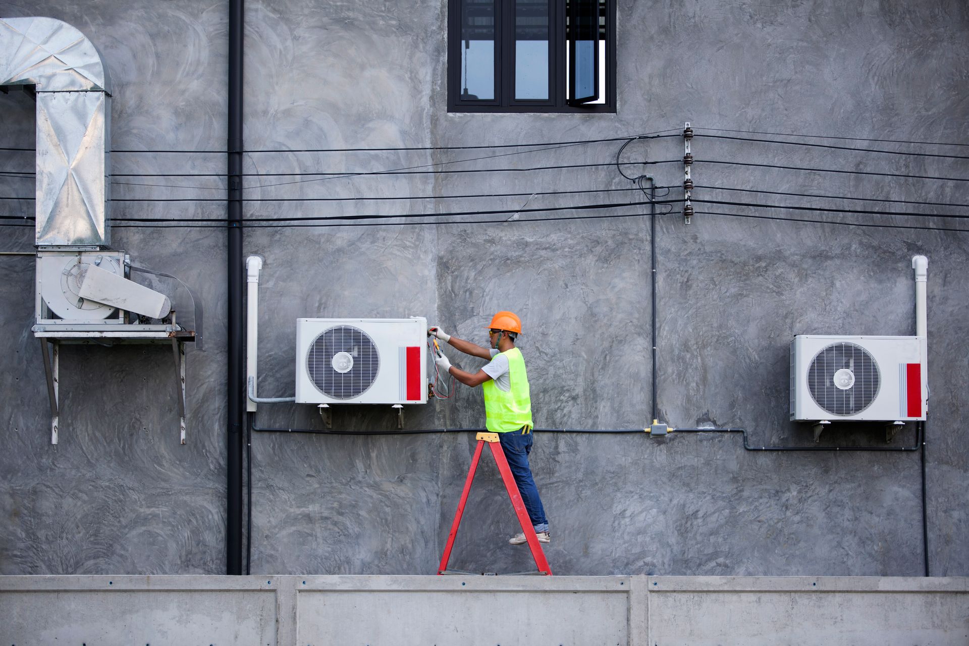 A man is standing on a ladder fixing air conditioners on the side of a building.