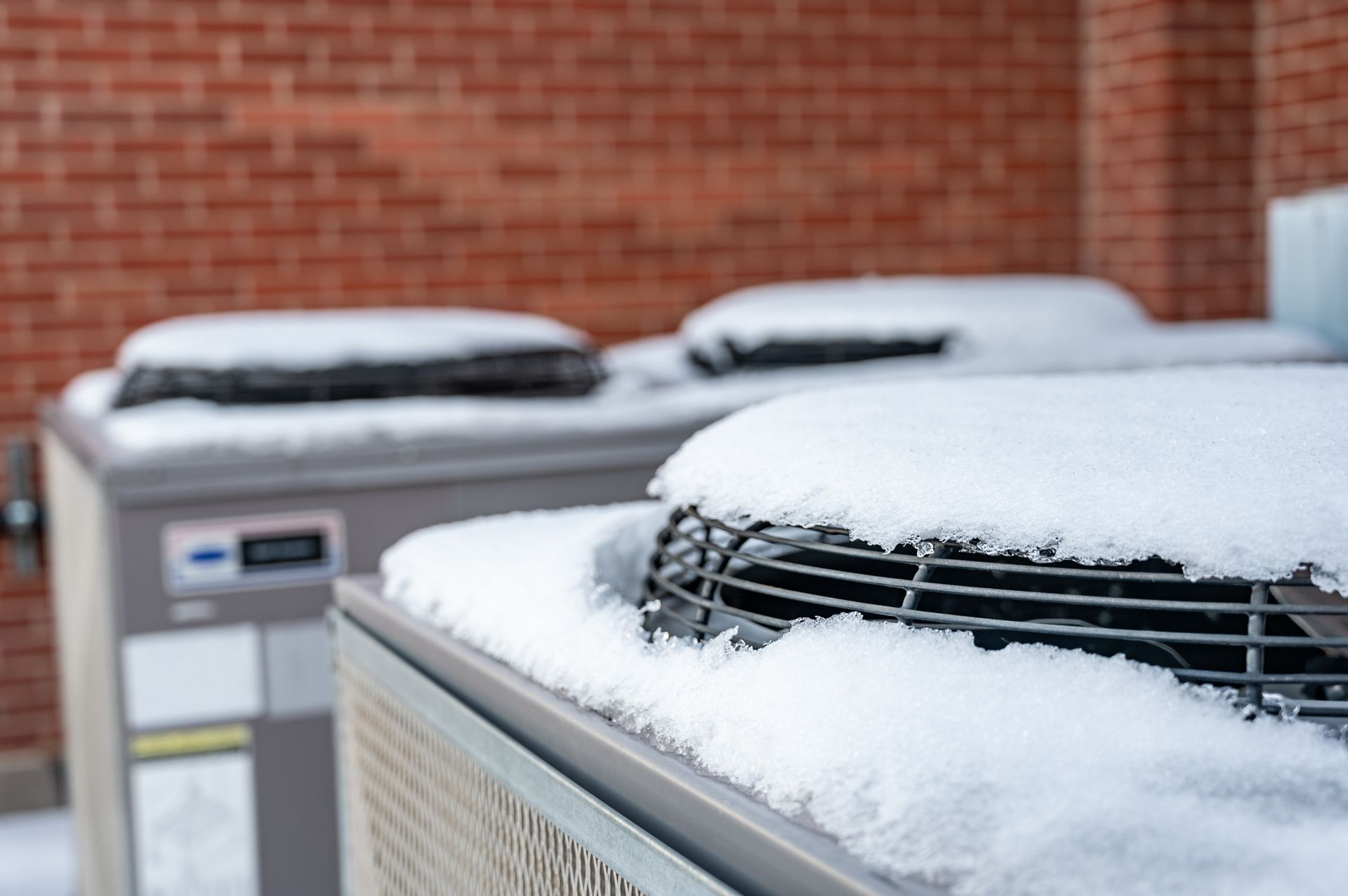 Two air conditioners are covered in snow in front of a brick building.