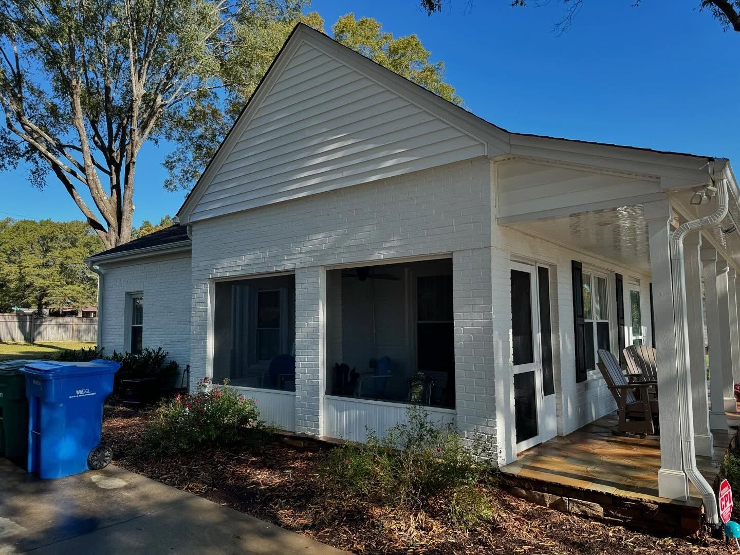 White brick house with screened porch and blue trash bins on a sunny day.