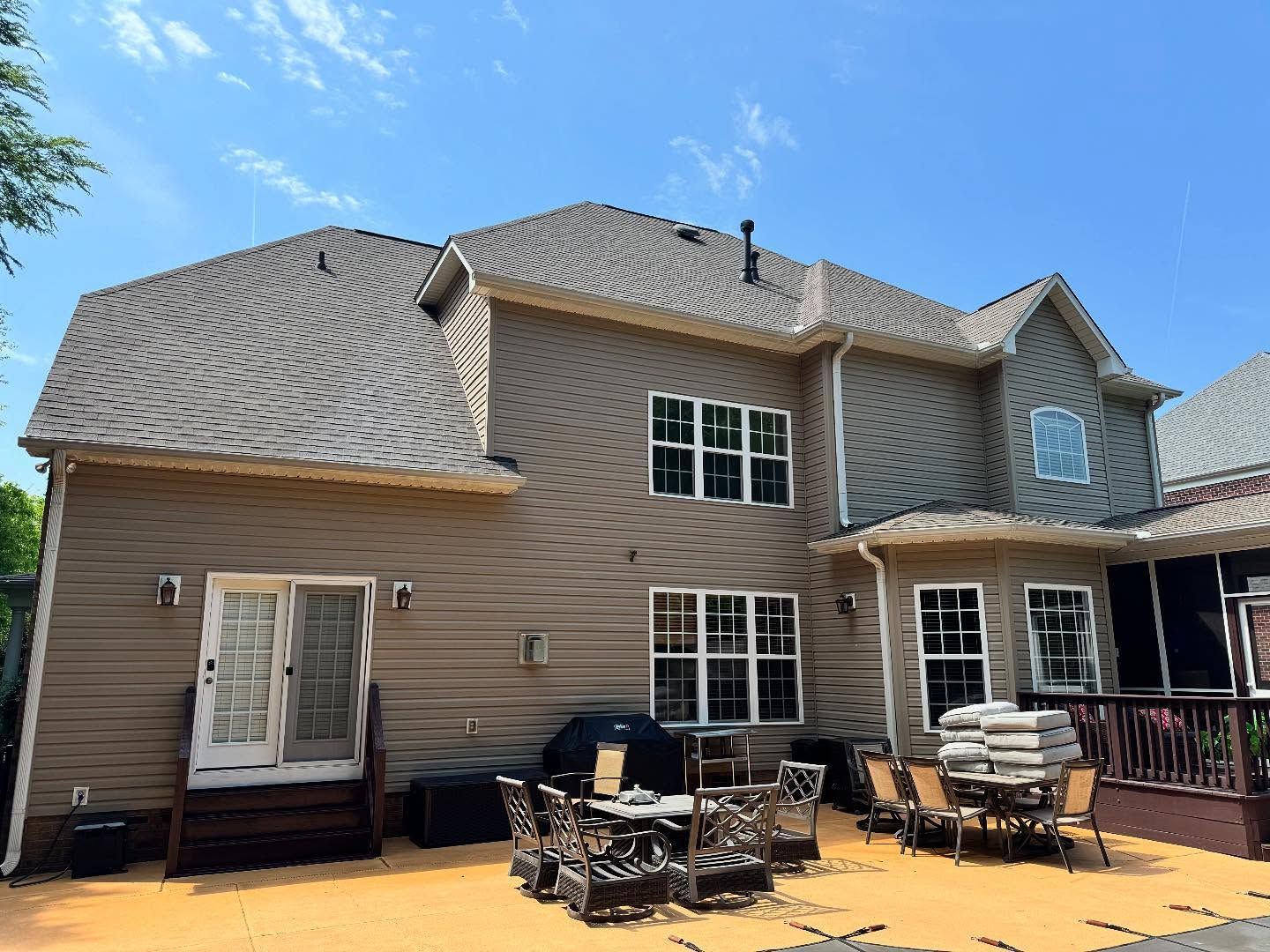 Back of a two-story brown house with a deck and patio. The house has a brown roof and tan siding, under a blue sky.