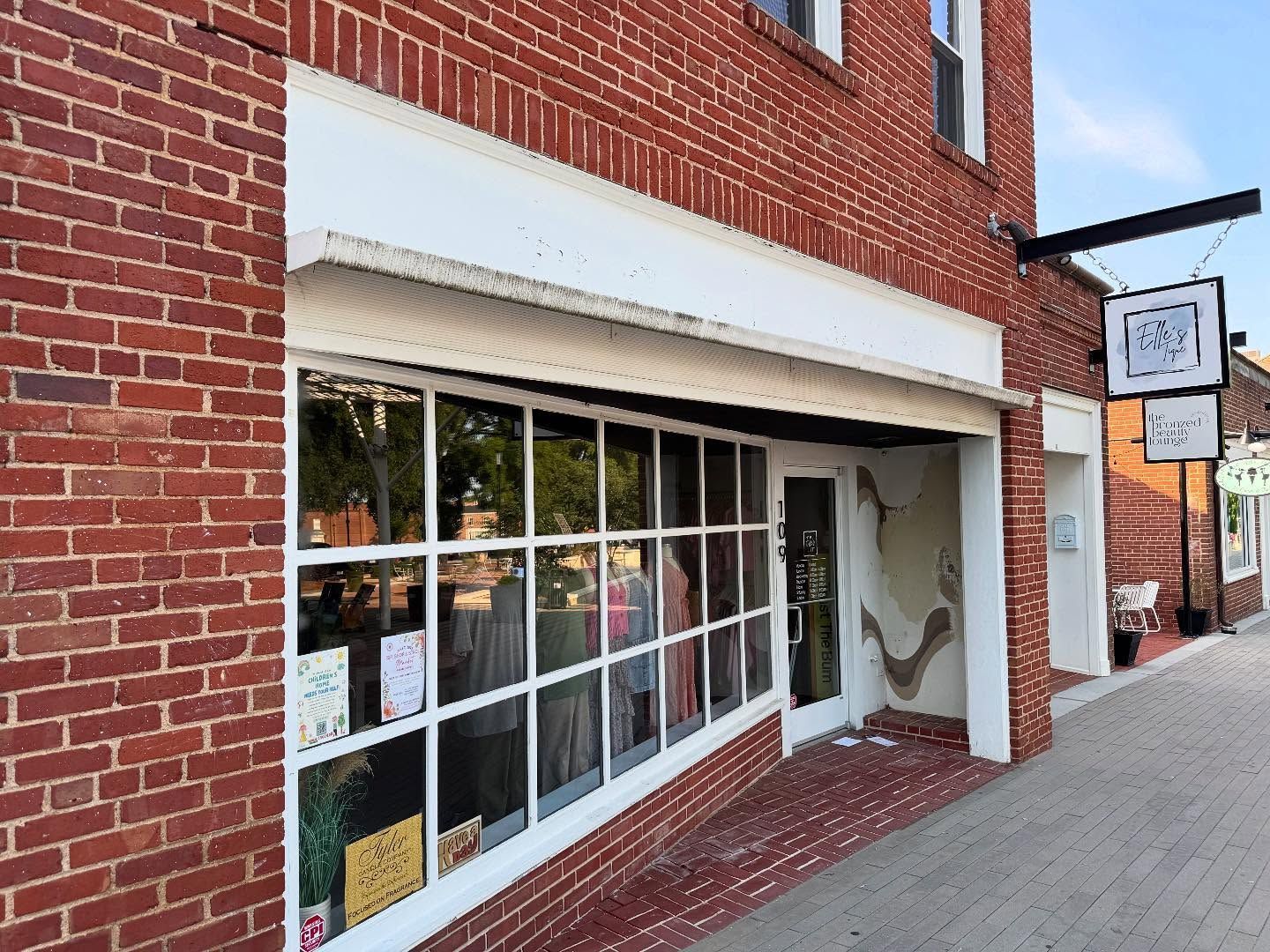 Red brick building with a shop front featuring large windows, white trim, and a sign. A door is slightly ajar.