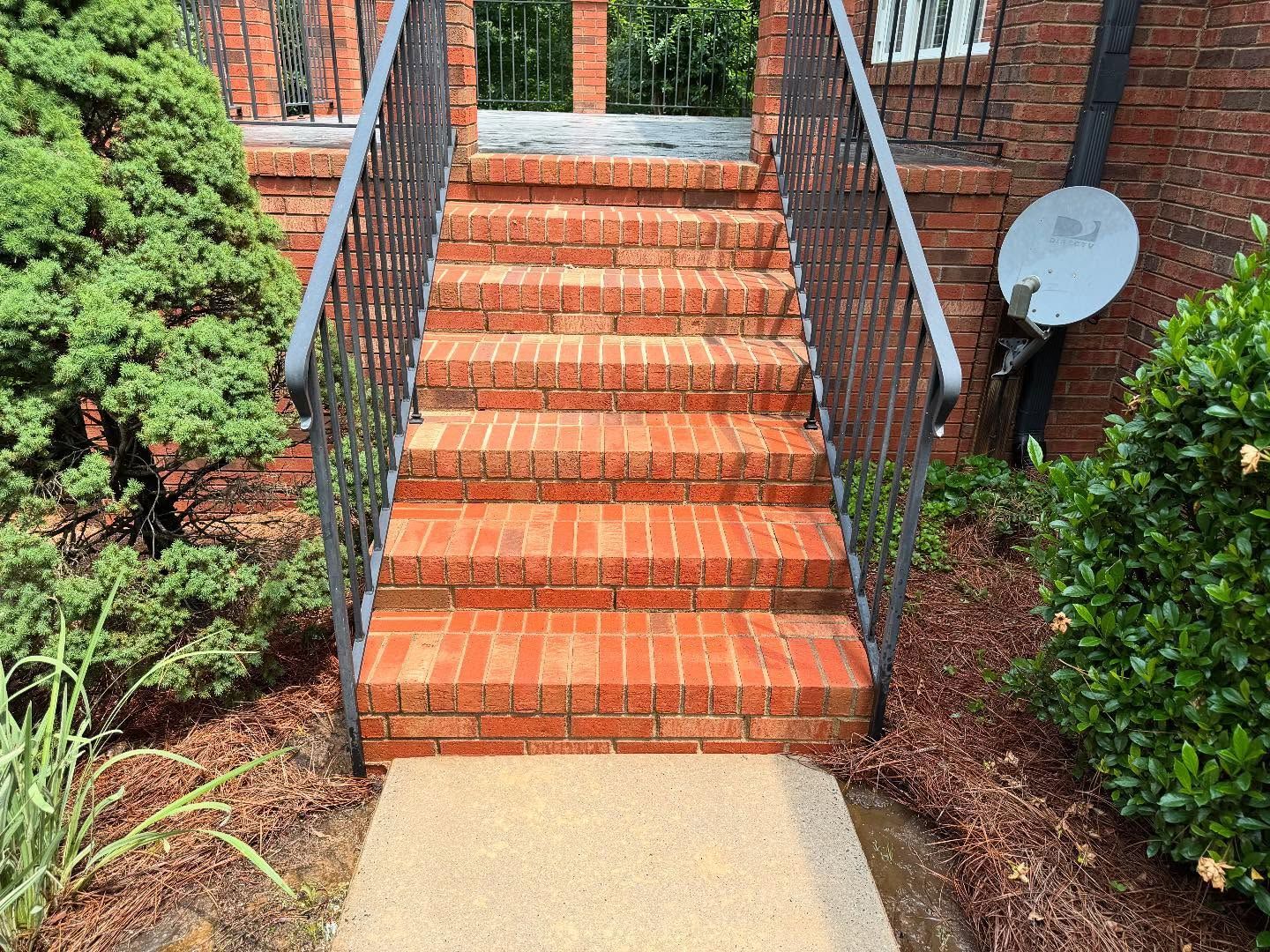Brick stairs with black metal railings leading up to a building entrance, flanked by green bushes and plants.