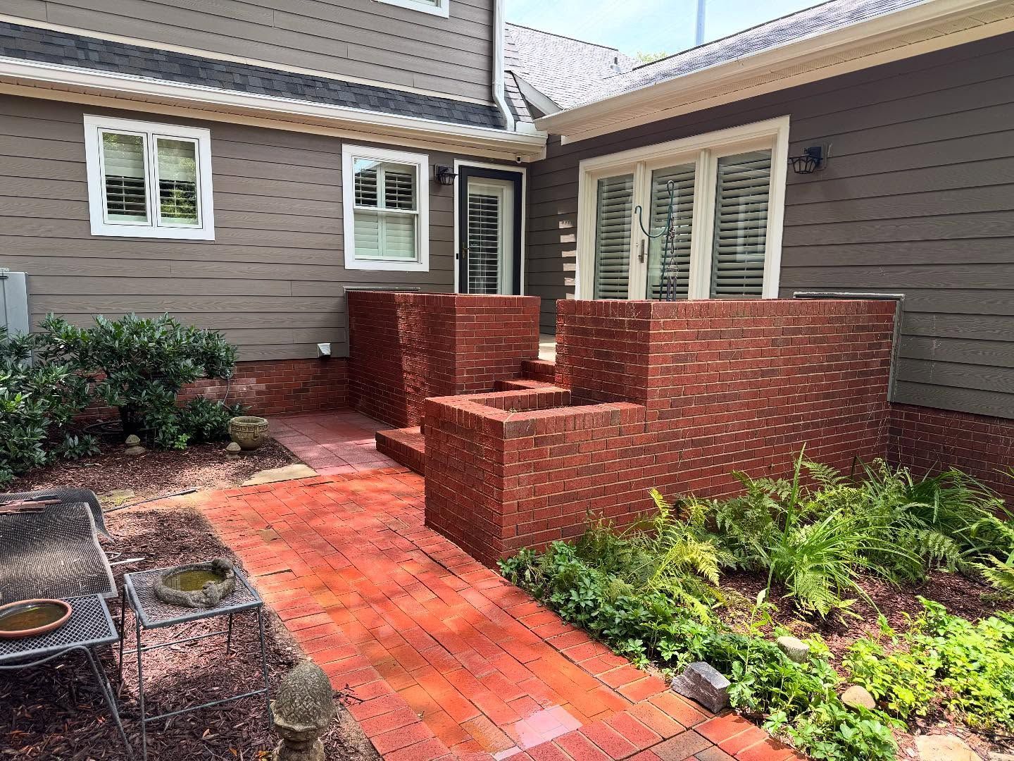 Brick patio with raised brick planter boxes and pathways, adjacent to a brown-sided house with windows and a door.