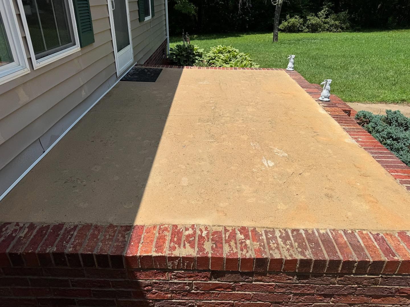 A concrete porch with brick edging in front of a tan house. Sunlight casts a shadow across the porch.