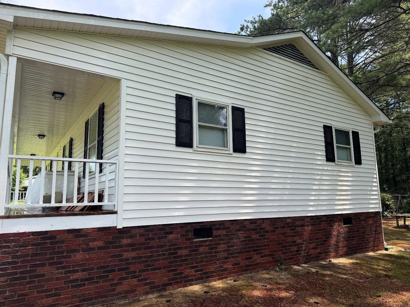 White-sided house with a red brick foundation, a porch with white railings, and black shutters on the windows.