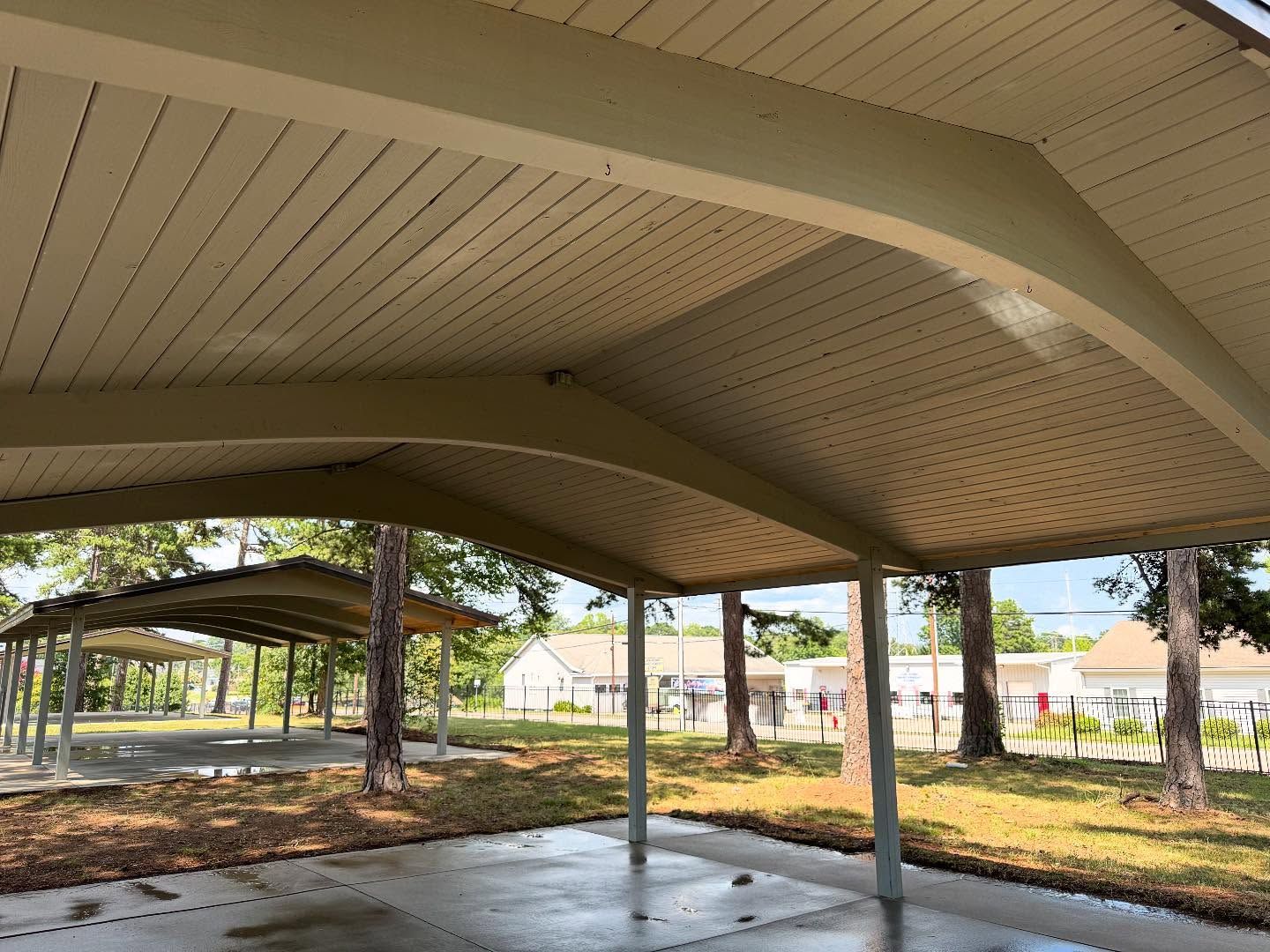 A covered outdoor structure with a curved roof. The ground is concrete, and trees are visible in the background.