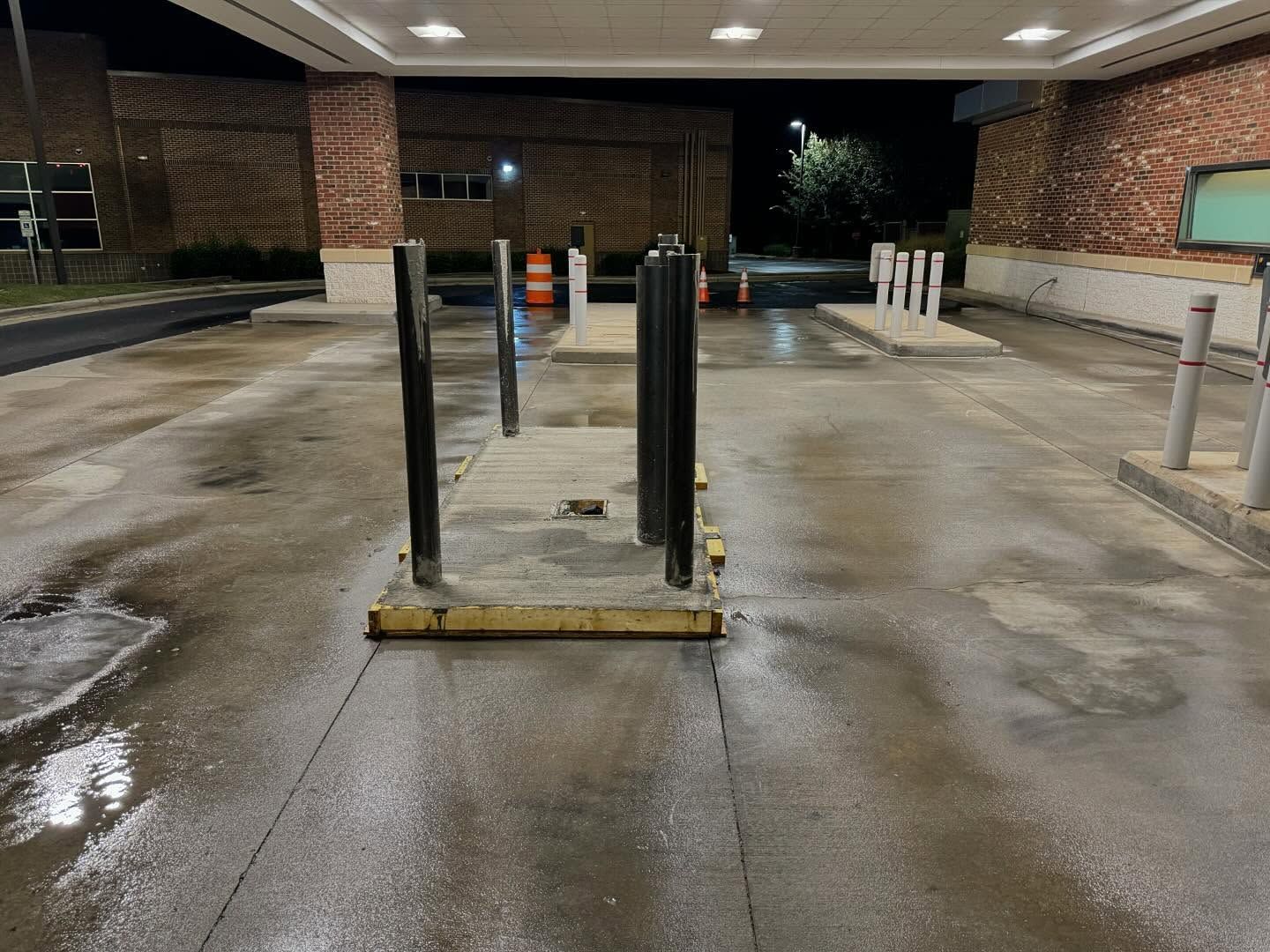 A wet, outdoor bank drive-through at night. Concrete and brick building with safety poles and lights.
