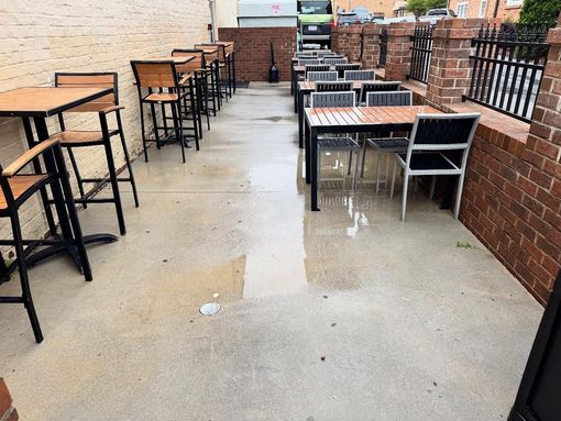 Outdoor restaurant seating area, wet concrete floor. Tables and chairs are empty, brick walls and a black iron fence visible.