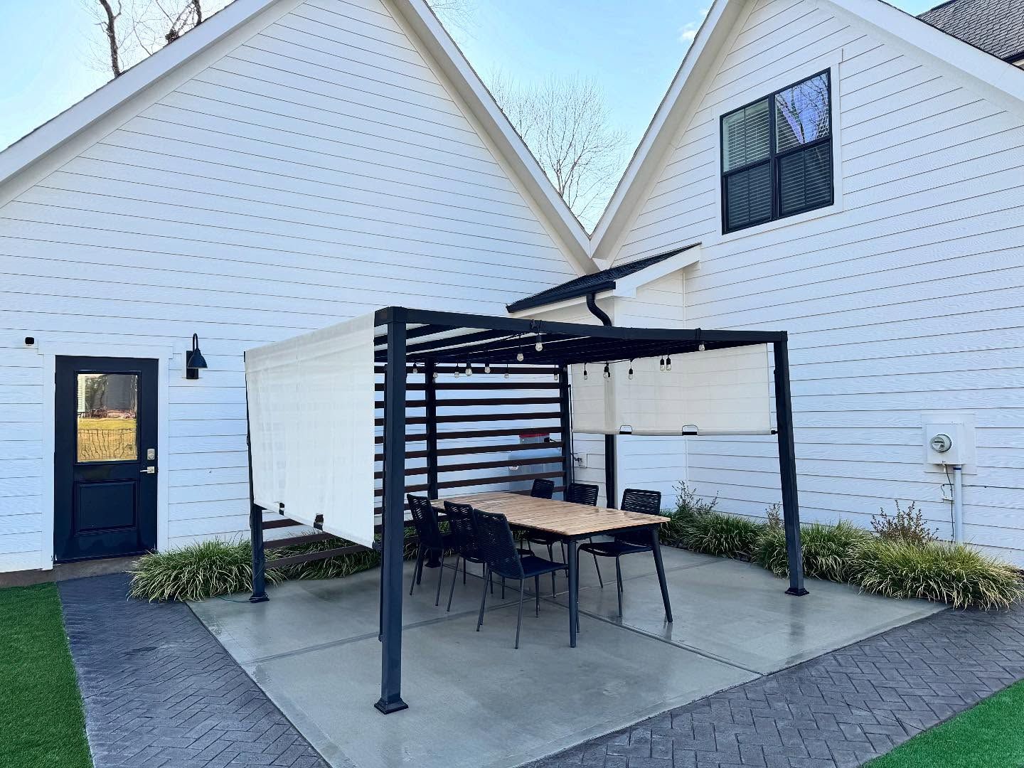 A modern outdoor dining area with a black pergola, a wooden table, and black chairs, situated between two white buildings.