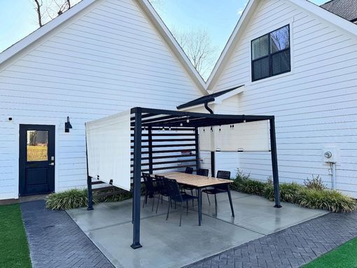 A modern outdoor dining area with a pergola, table, and chairs, set between two white buildings.